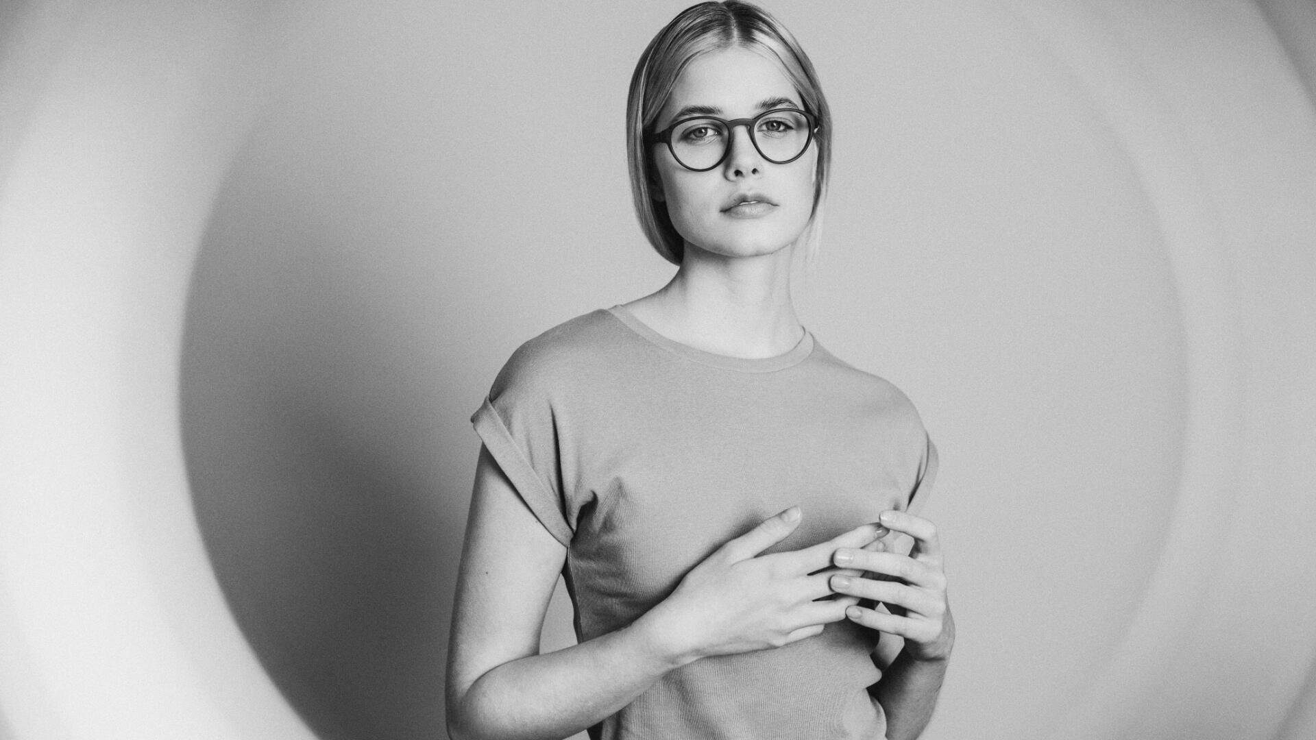 Schwarzweißfoto einer jungen Frau mit hellem Haar, runder Brille und schlichtem T-Shirt, die vor einem sanft beleuchteten Hintergrund steht und ihre Hände sanft ihre Brust berühren.