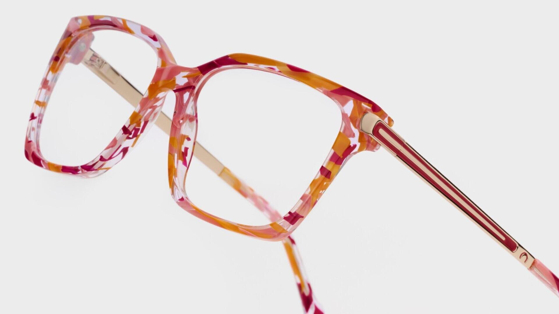 Close-up of colorful eyeglasses with a square frame, featuring a marbled pattern in shades of pink, orange, and white, and gold metal arms, set against a plain white background.
