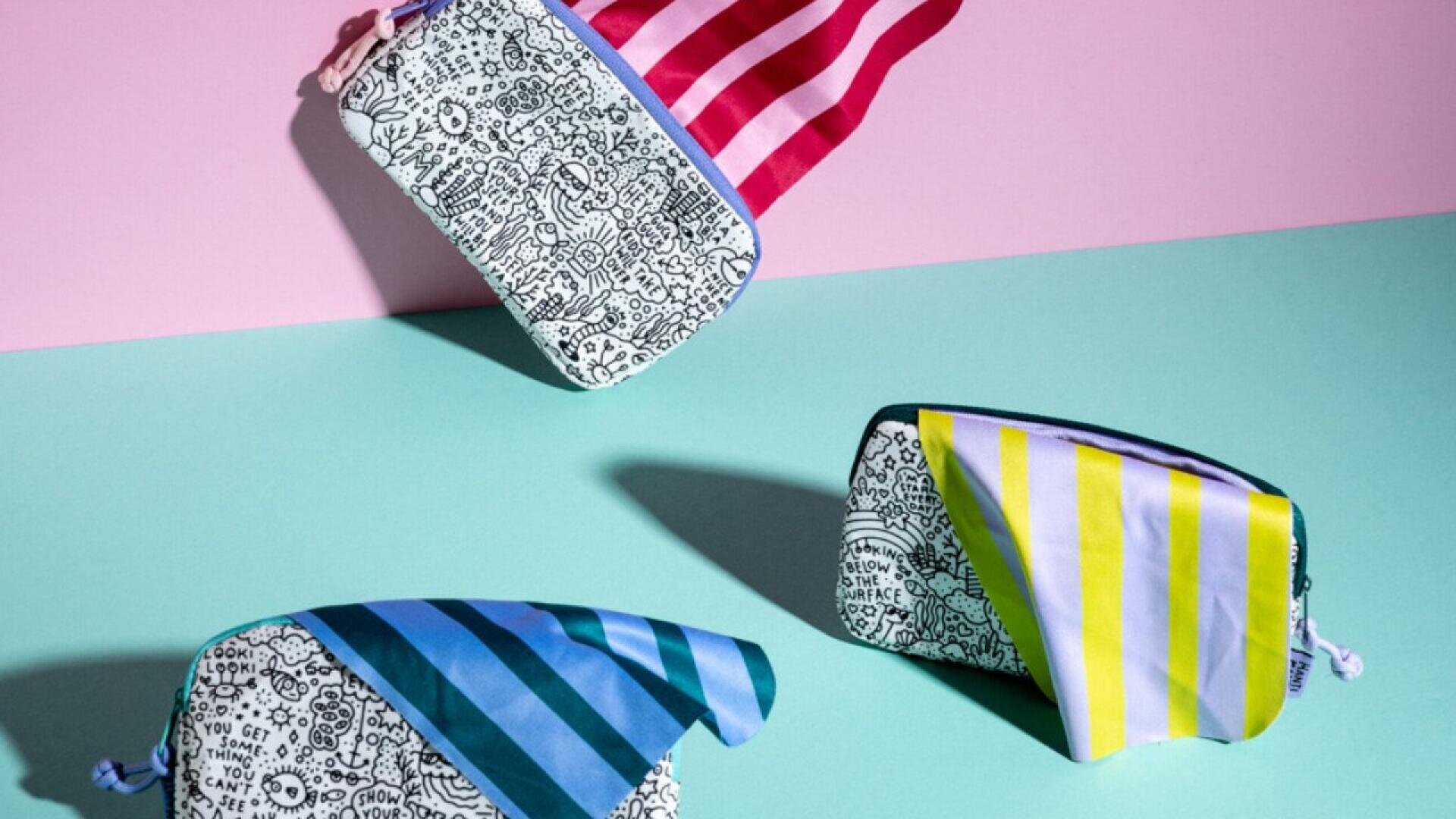 Three patterned pouches with black-and-white doodle designs and bold striped flaps (pink, blue, and yellow) are displayed on a pastel pink and teal background, with one pouch appearing to float in the air.
