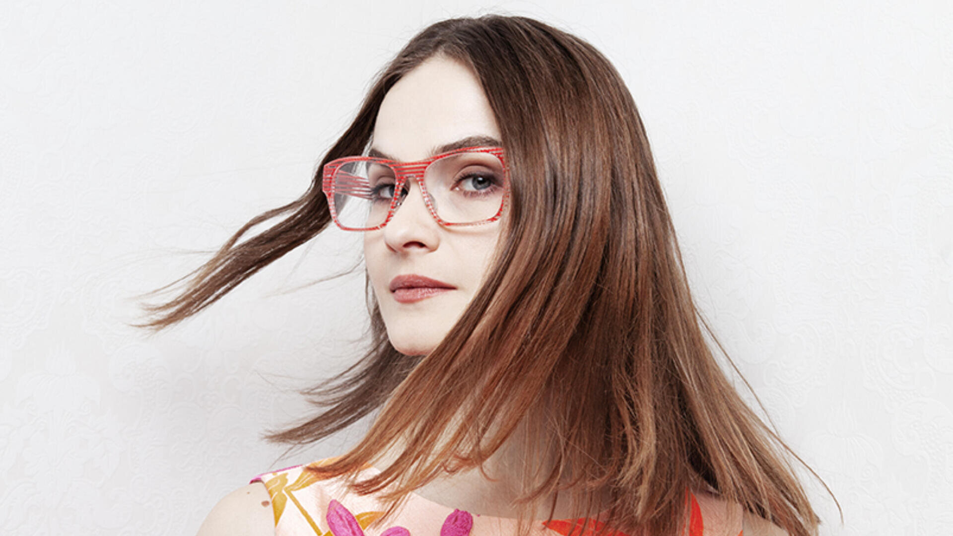 Eine Frau mit glattem braunem Haar, einer Brille mit rotem Rahmen und einem bunten Oberteil mit Blumenmuster blickt in die Kamera. Ihr Haar fällt ihr vor einem schlichten weißen Hintergrund teilweise ins Gesicht.