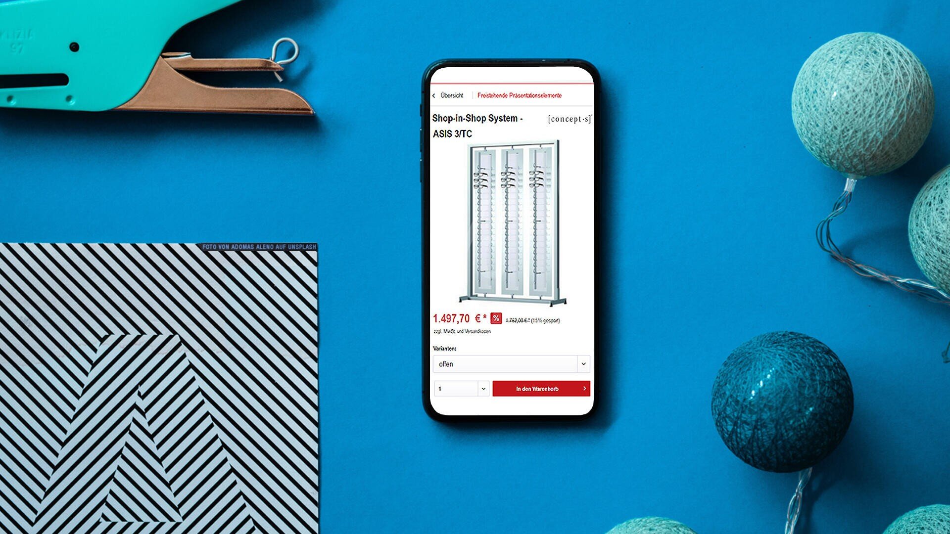 Ein Smartphone auf einer blauen Fläche zeigt eine Onlineshop-Seite für ein Regal an. In der Nähe befinden sich ein Hefter, ein schwarz-weiß gestreiftes Notizbuch und eine dekorative Lichterkette mit blauen und weißen Kugeln.
