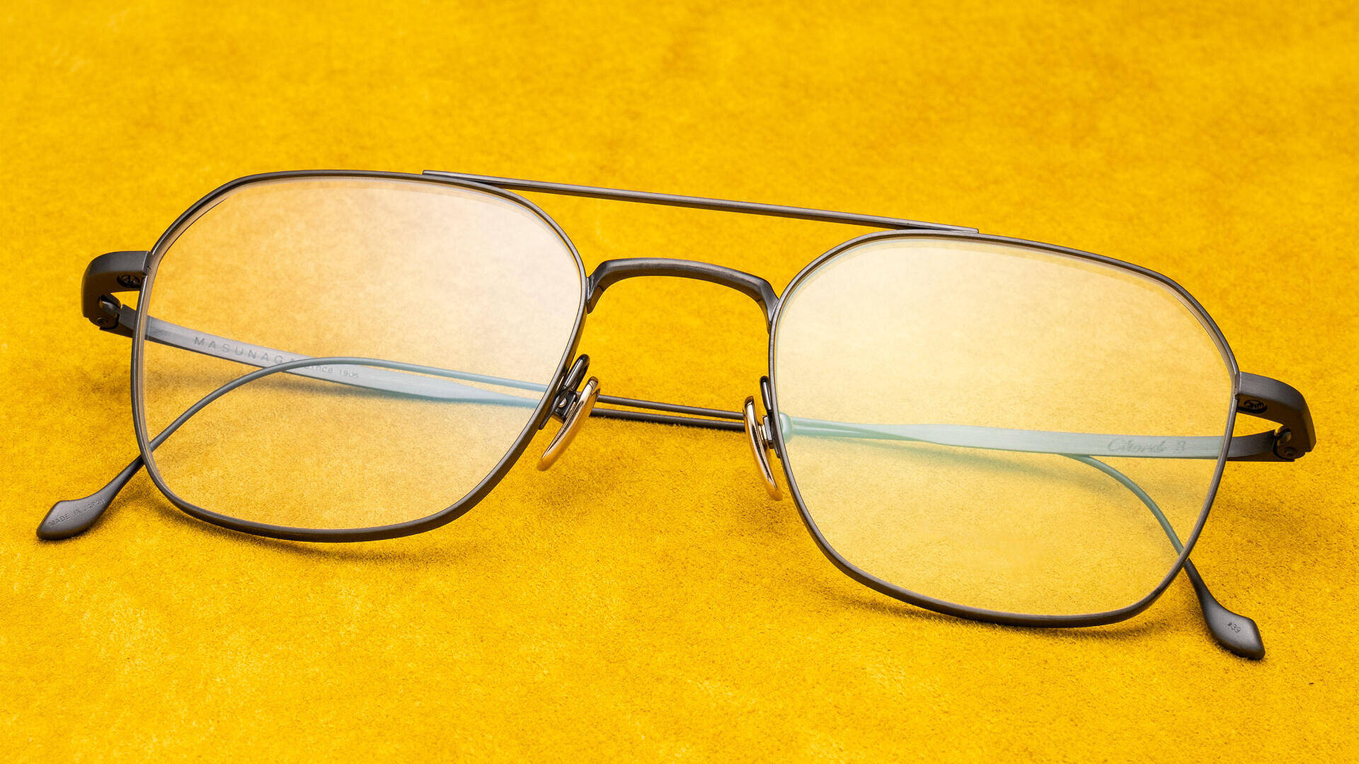 Eine Brille mit Metallrahmen und gelb getönten Gläsern liegt auf einer leuchtend gelben strukturierten Oberfläche.