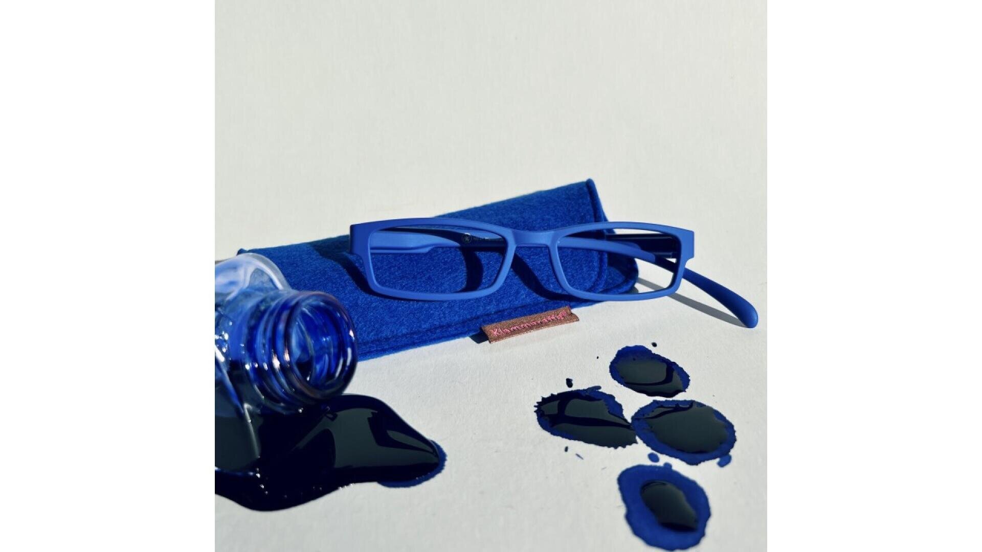 Bright blue eyeglasses rest on a matching case beside a tipped-over ink bottle, with several pools of spilled dark blue ink on a light surface.