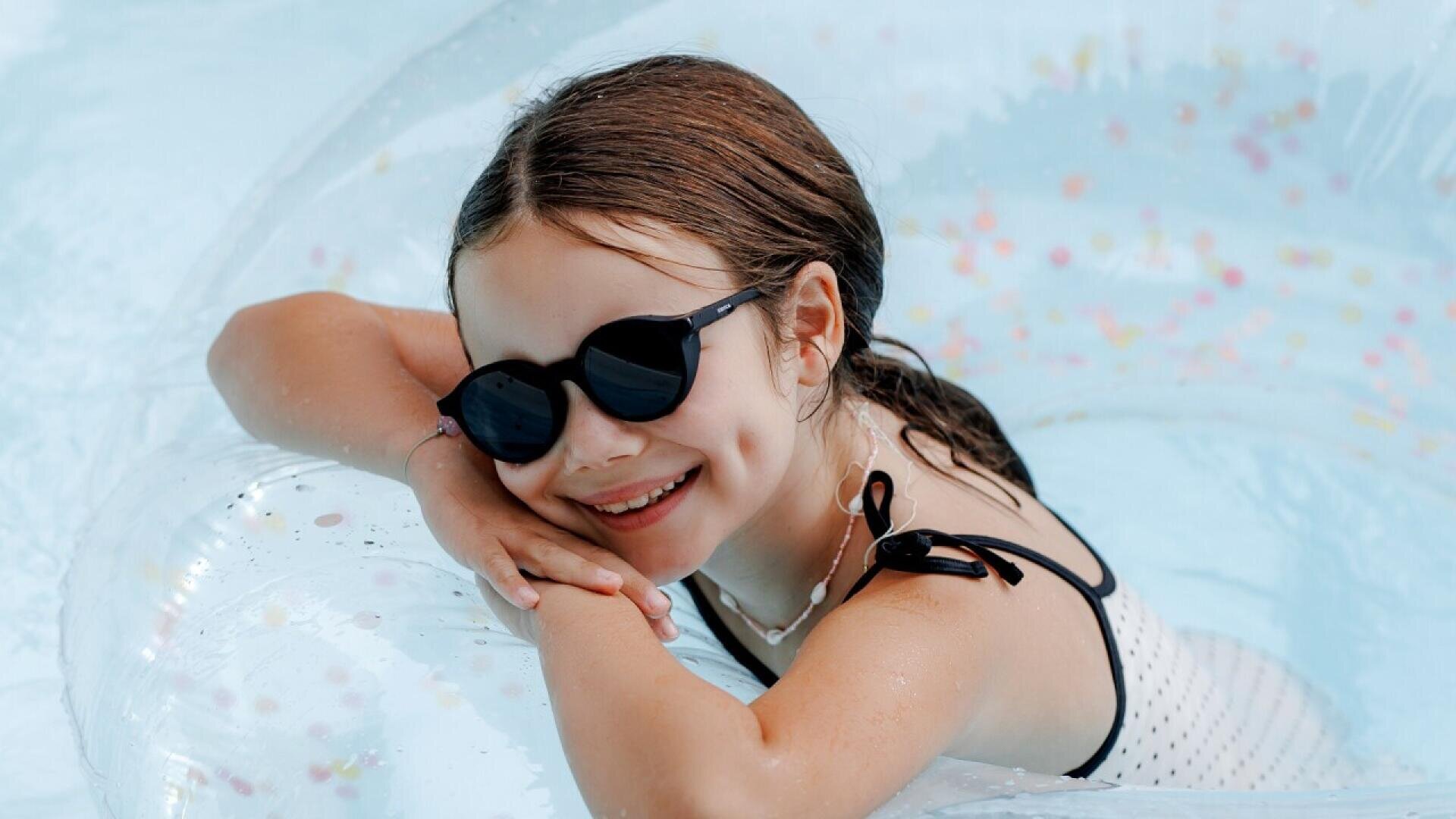 A smiling young girl wearing black sunglasses and a white swimsuit with black straps leans on a clear inflatable float in a pool. Her hair is wet and she appears happy and relaxed in the water.
