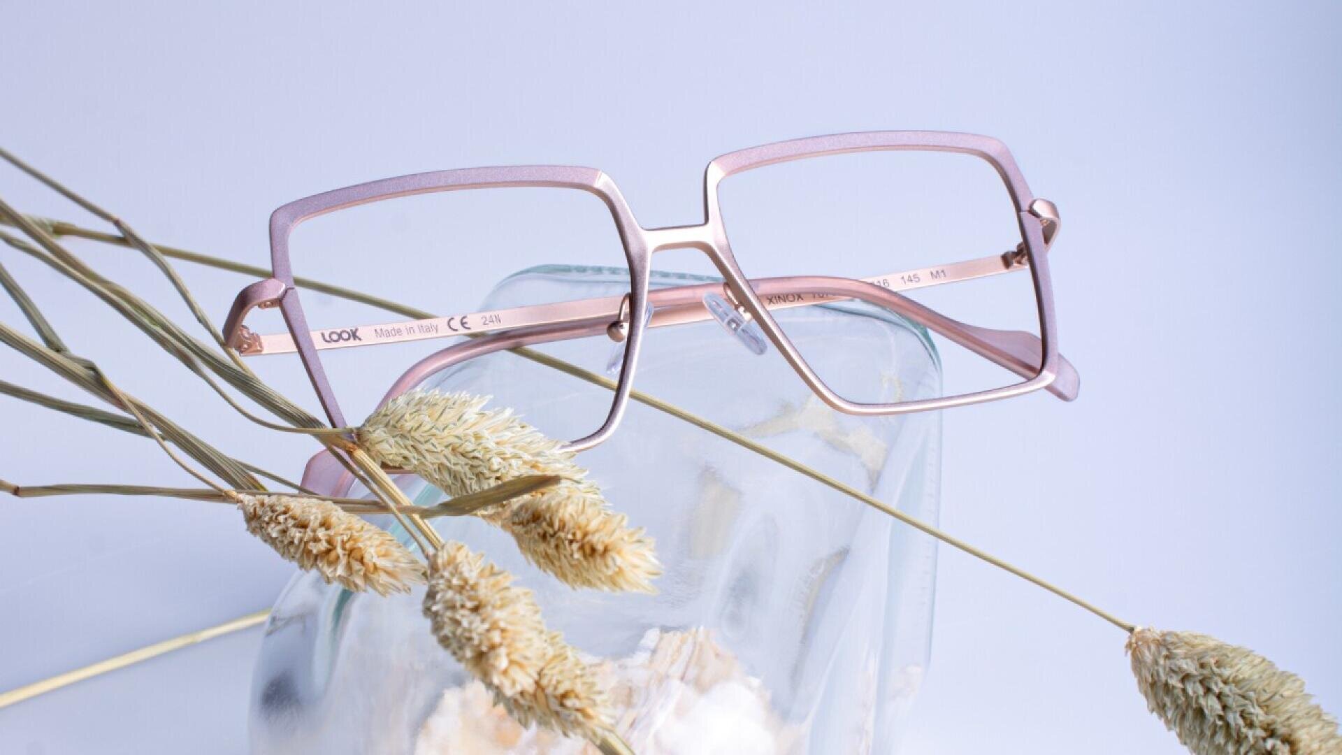 Eine stilvolle, quadratische, rosa Brille liegt auf einem durchsichtigen Glasgefäß, umgeben von beigen, getrockneten Gräsern vor einem hellen Pastellhintergrund.