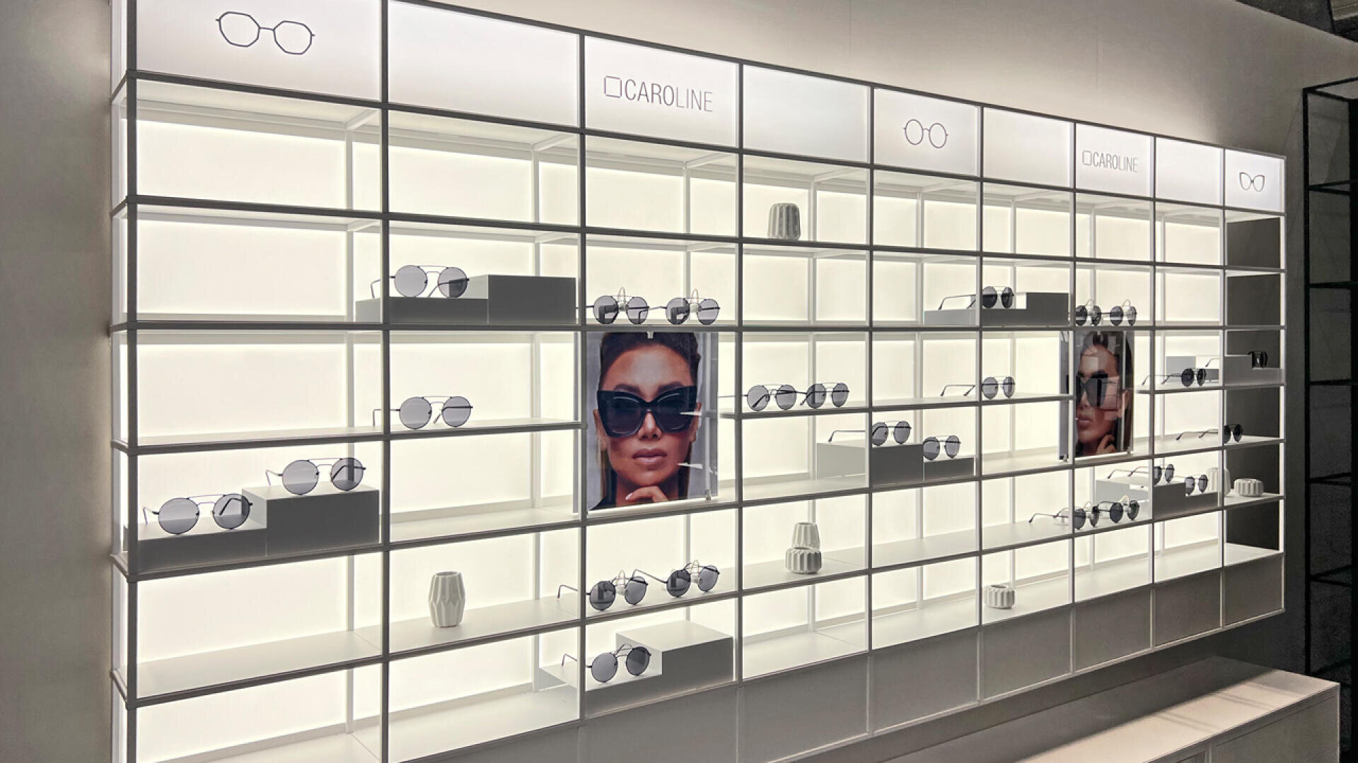 Eine moderne, gut beleuchtete Brillenwand mit Regalen, in denen verschiedene Sonnenbrillen und Brillen präsentiert werden. Es gibt zwei große Fotos einer Frau, die eine Sonnenbrille trägt, und der Markenname CAROLINE ist auf beleuchteten Schildern zu sehen.