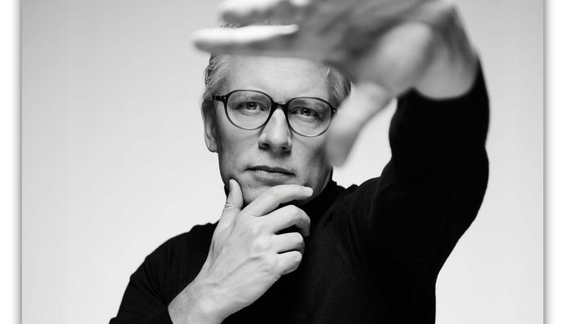 Schwarzweißfoto einer Person mit Brille, die nachdenklich in die Kamera blickt, eine Hand am Kinn und die andere nach vorne ausgestreckt, sodass ihr Gesicht teilweise verdeckt ist.