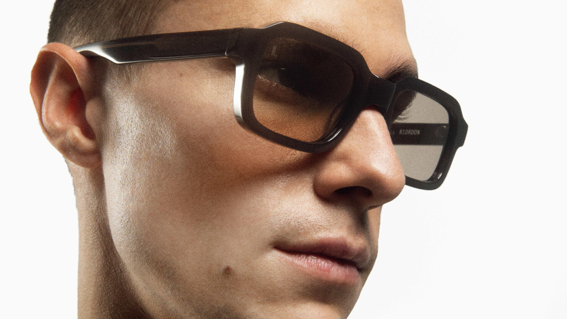 Close-up of a person wearing dark, geometric-shaped sunglasses with a translucent brown tint, looking slightly to the side against a plain white background.