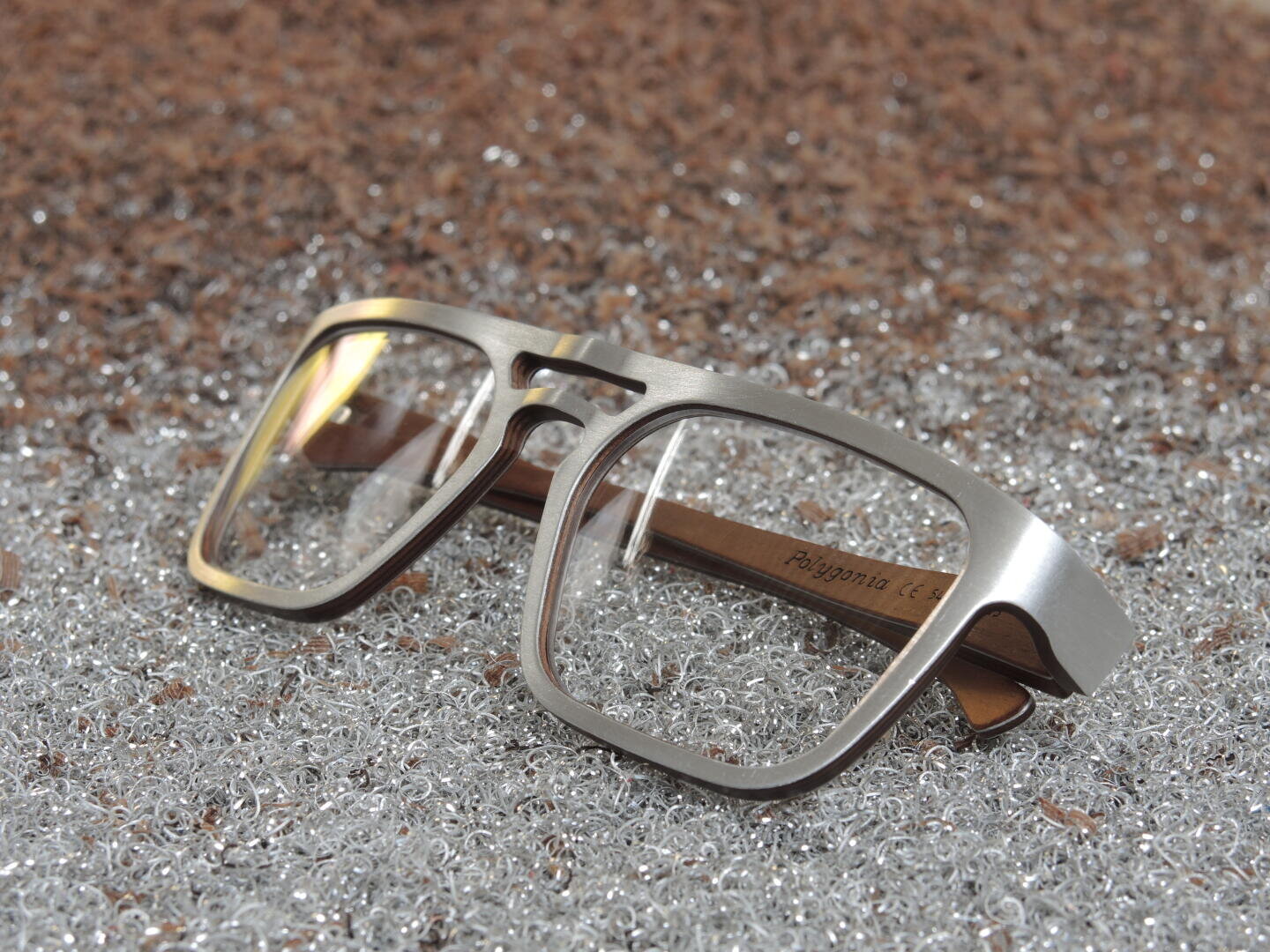 Eine metallgerahmte Brille mit rechteckigen Gläsern und Holzbügeln ruht auf einer glitzernden, strukturierten Silberoberfläche.