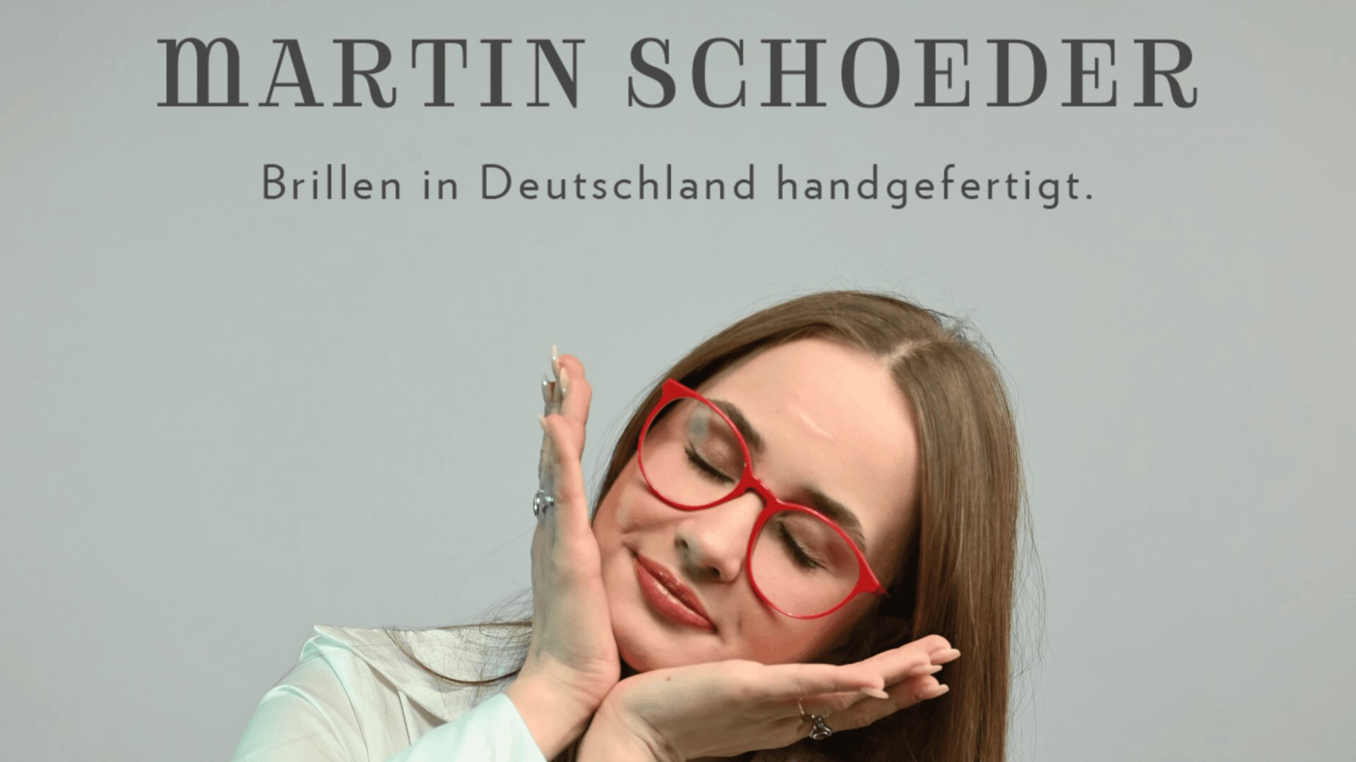 Eine Frau mit langen braunen Haaren und roter Brille lächelt mit geschlossenen Augen und stützt ihr Gesicht auf ihre Hände. Oben steht: Martin Schoeder. Brillen in Deutschland handgefertigt.