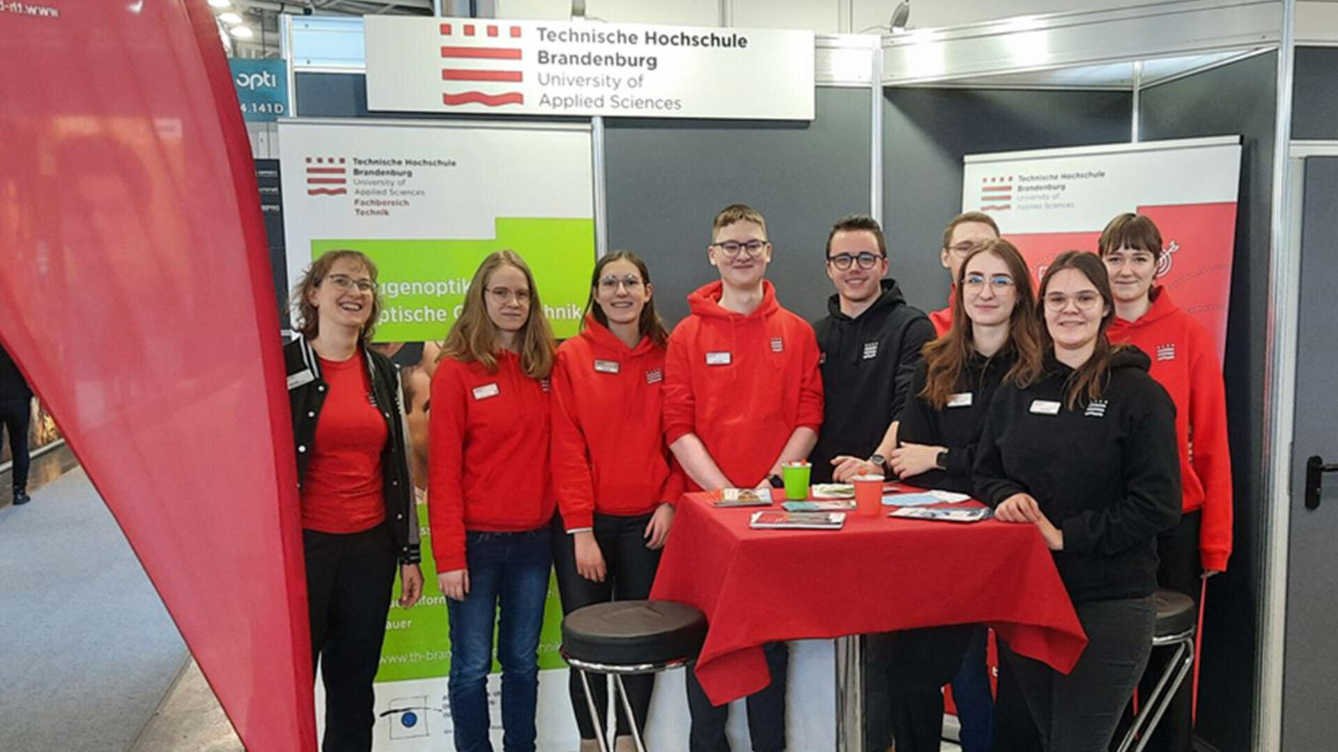 Eine Gruppe von neun Studierenden, die meisten in rot-schwarzen Kapuzenpullovern, posiert lächelnd an einem Stand der Technischen Hochschule Brandenburg. Sie stehen hinter einem hohen roten Tisch mit Werbematerialien.