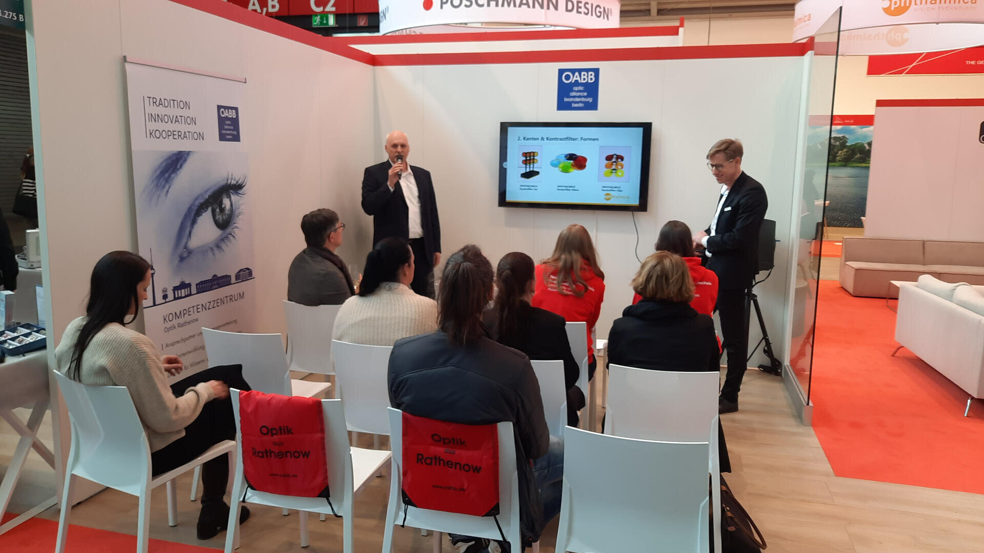 A group of people sit facing a presentation, where a man stands and speaks next to a screen displaying a colorful slide. Another man stands nearby. Red bags labeled “Optik Rathenow” hang on some chairs in the booth.
