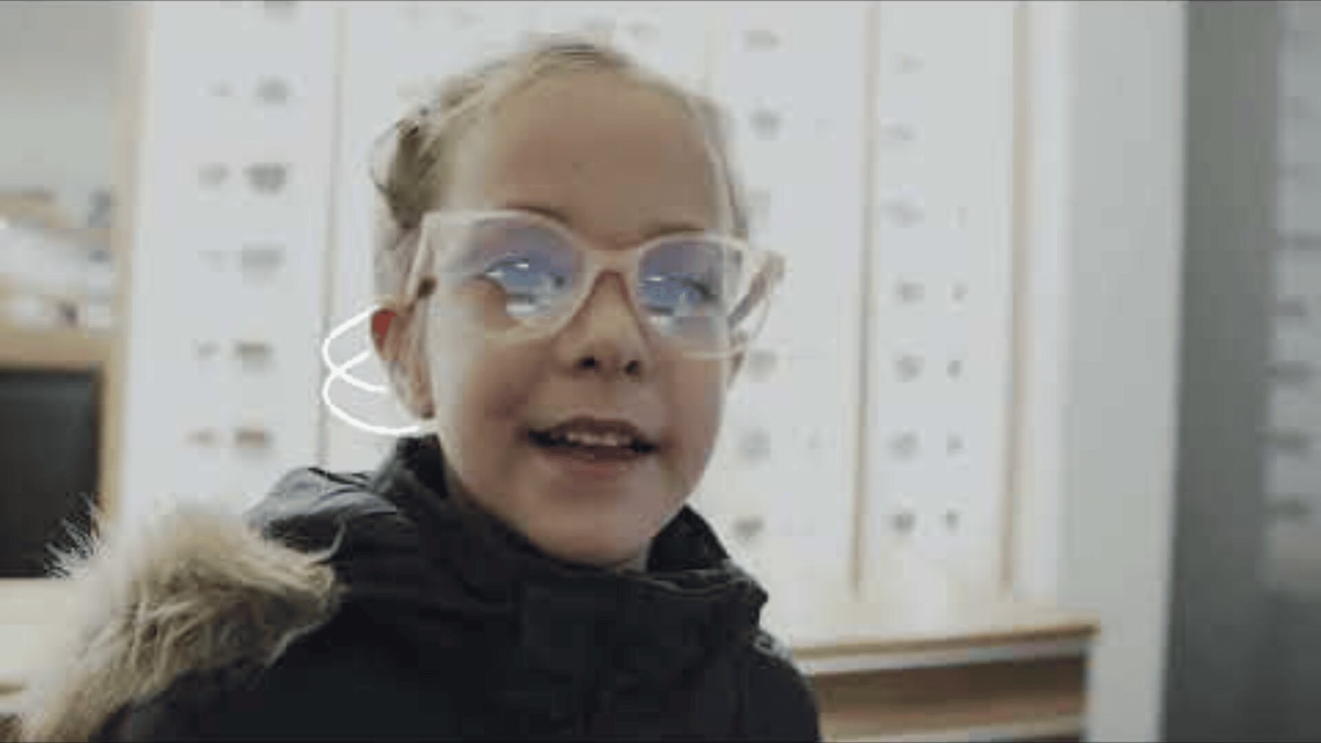 A young girl wearing large, see-through glasses and a dark coat with a fur-lined hood smiles indoors, with rows of glasses frames visible against a blurred background.