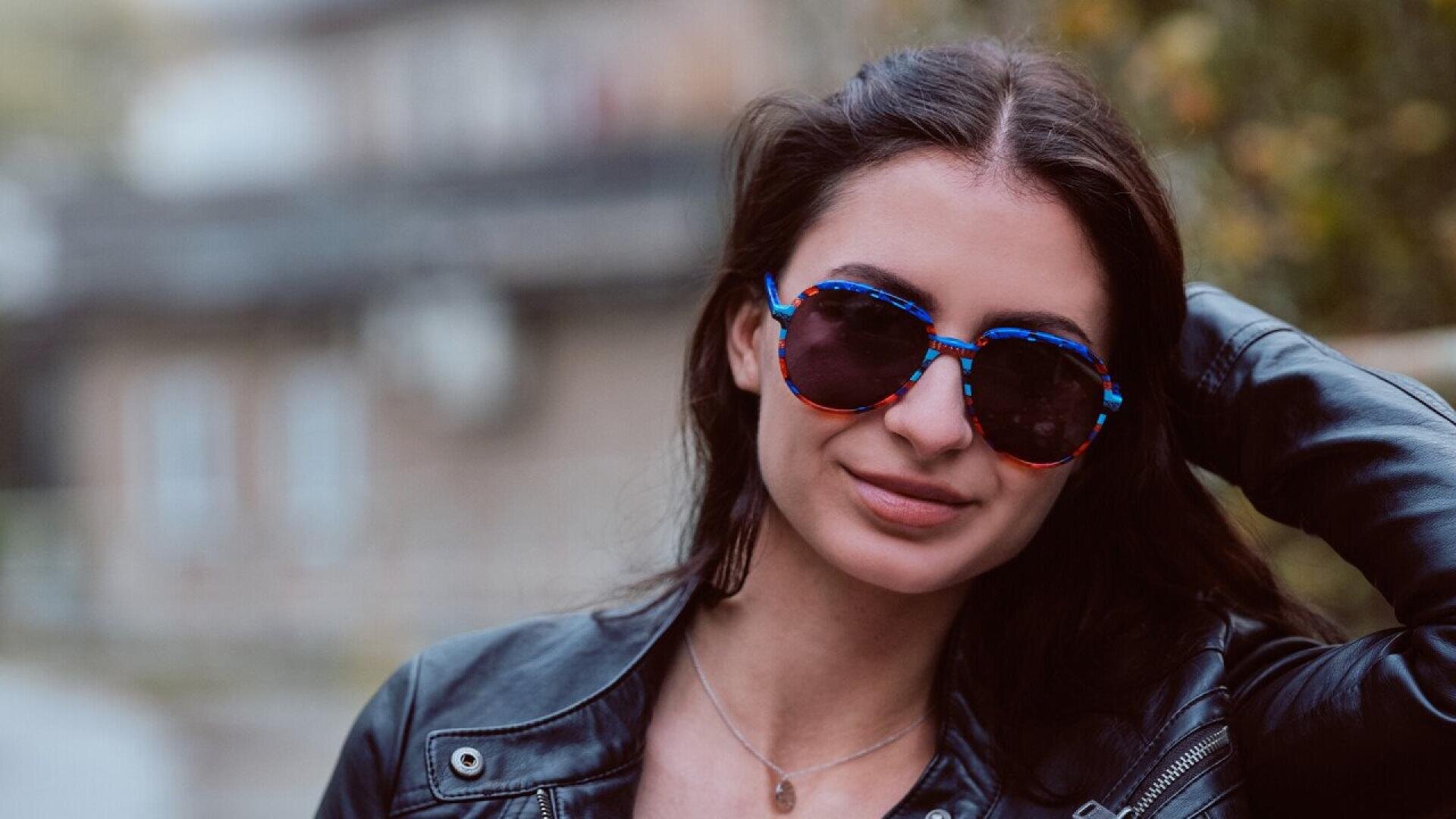 A woman with long dark hair wearing a black leather jacket and round blue-rimmed sunglasses smiles outdoors, with one hand touching her head and a blurred background behind her.