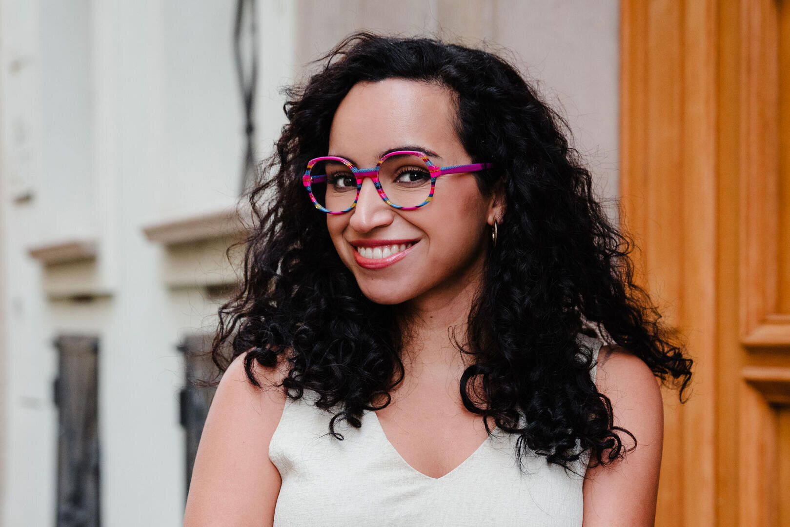 Eine Frau mit lockigem dunklem Haar, einer rosa Brille und einem ärmellosen hellen Top lächelt, während sie draußen in der Nähe eines beigen Gebäudes mit Holz- und Steindetails steht.