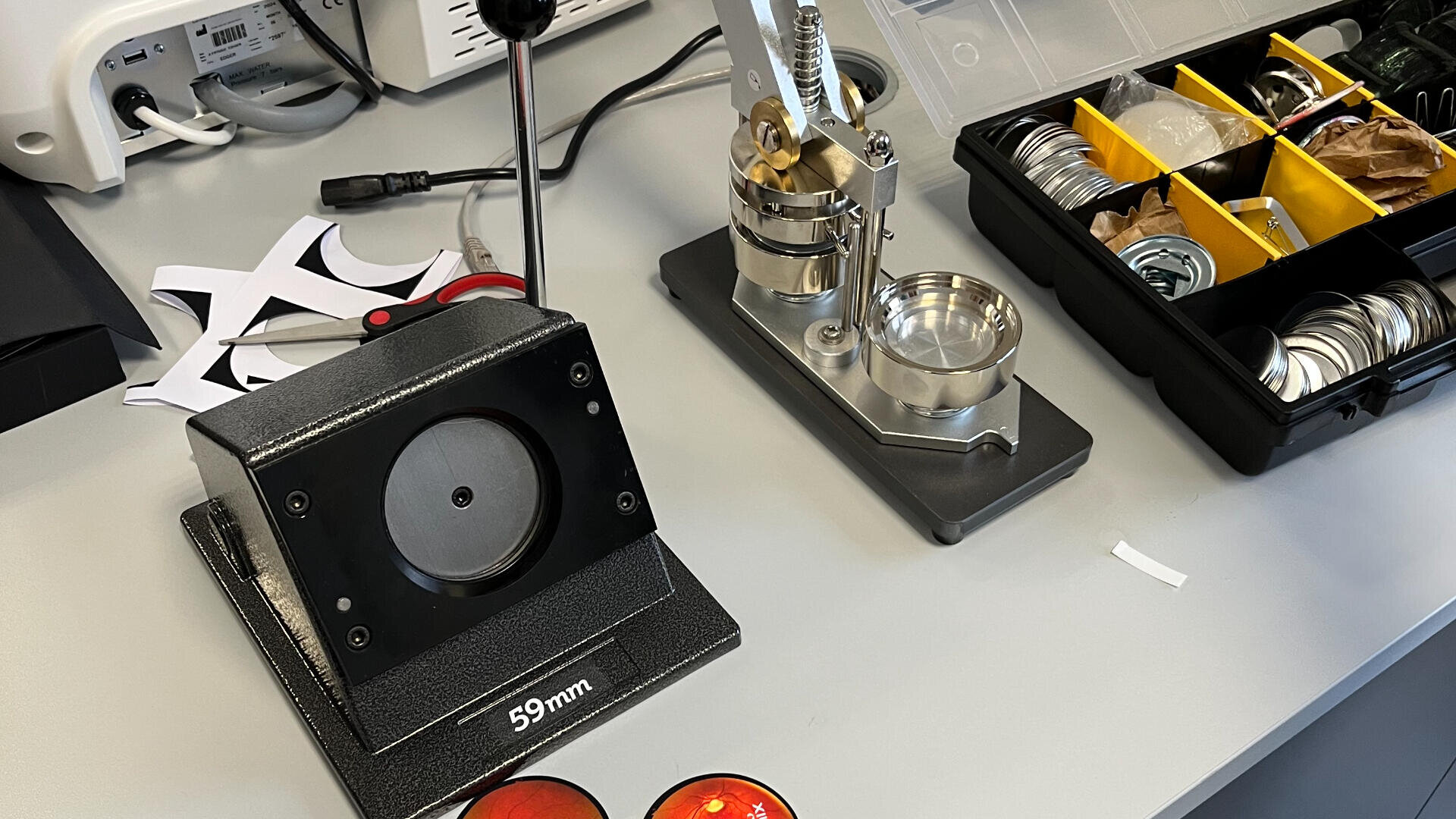 Eine 59-mm-Maschine zur Herstellung von Buttons auf einem Schreibtisch. In den Fächern daneben sind Buttonteile, Werkzeuge und eine Kiste mit Zubehör verstaut.