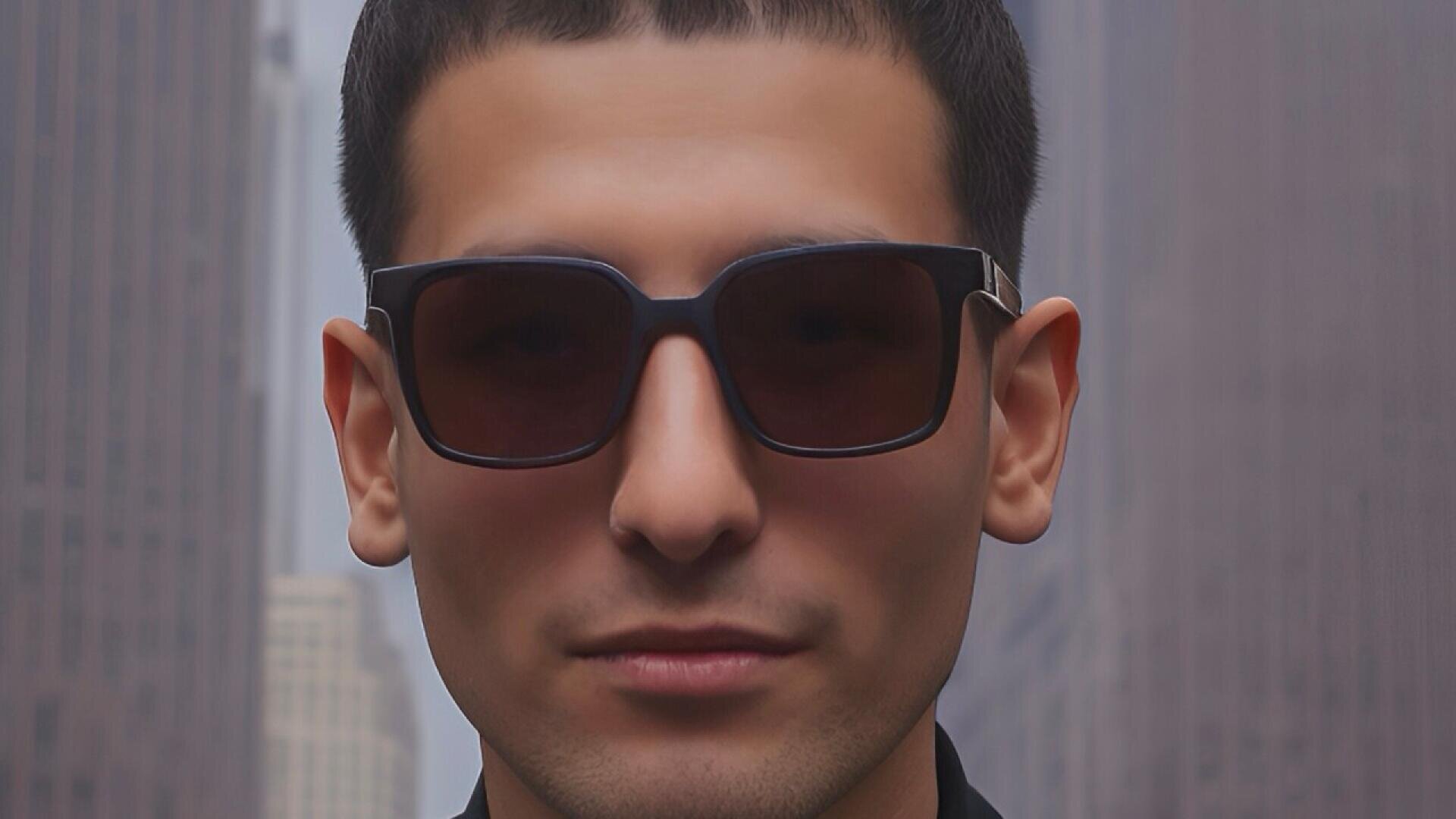 A man with short dark hair and wearing black sunglasses stands outdoors in a city with tall, blurred buildings in the background.