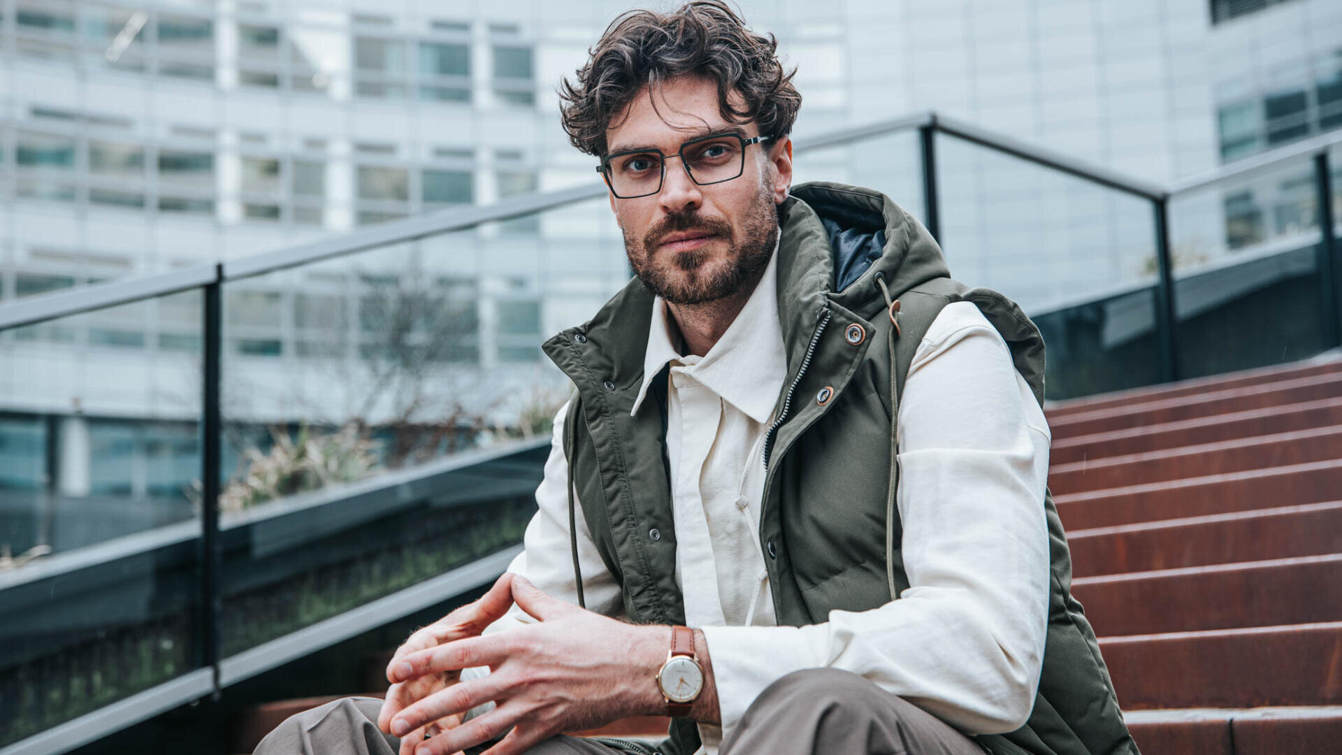 Ein Mann mit lockigem Haar, Bart und Brille sitzt auf einer Freitreppe. Er trägt ein weißes Hemd, eine olivgrüne Weste, eine braune Hose und eine Armbanduhr. Im Hintergrund sind moderne Glasgebäude zu sehen.