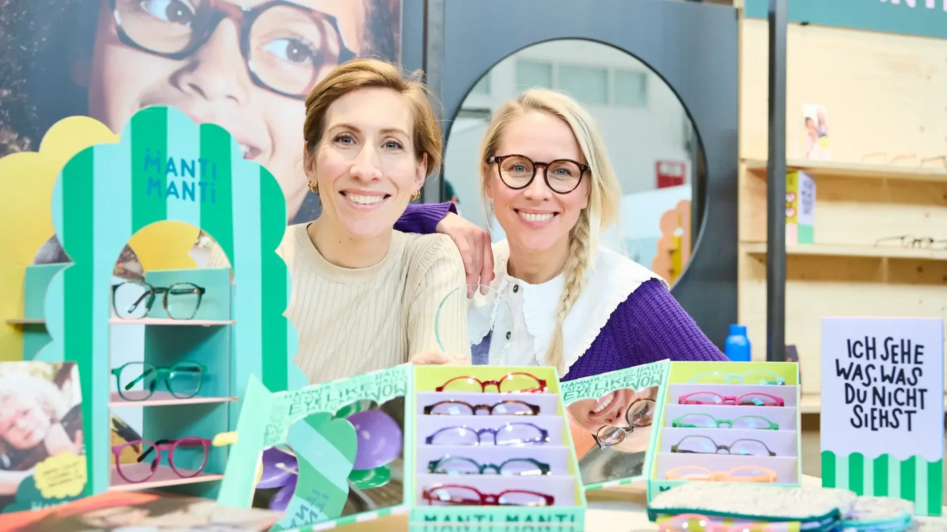 Two smiling women with glasses stand behind a display of colorful eyeglasses. One wears a beige top, the other a purple sweater. Signs and boxes with glasses are on the table, with a German sign reading Ich sehe was, was du nicht siehst.