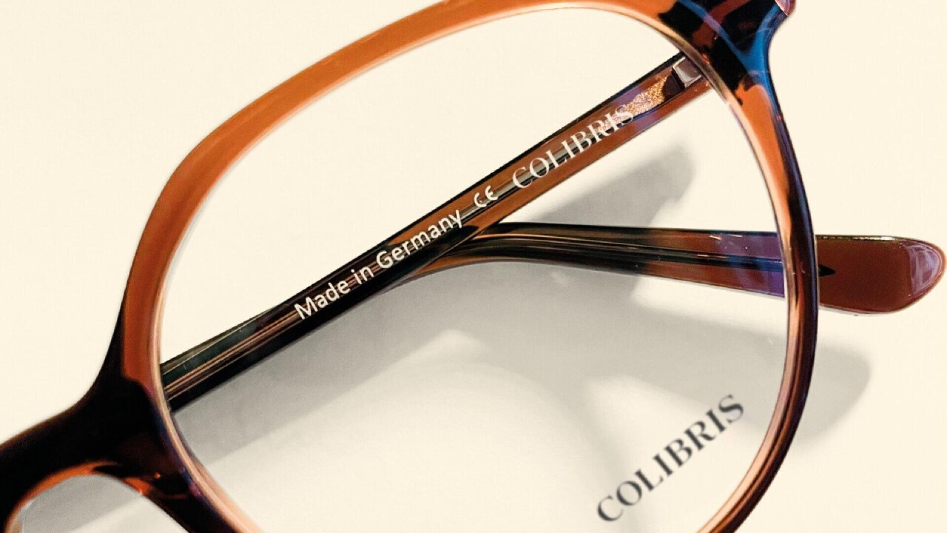 Close-up of brown tortoiseshell eyeglasses showing the text Made in Germany CE COLIBRIS on the inner arm, with COLIBRIS reflected on a white surface below.
