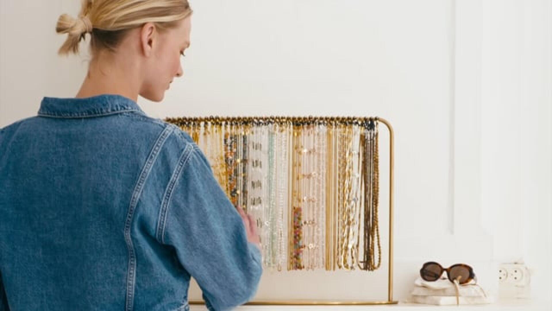 A woman in a denim jacket stands in front of a jewelry stand with colorful necklaces and bracelets. Towels and sunglasses lie on the white counter next to her.