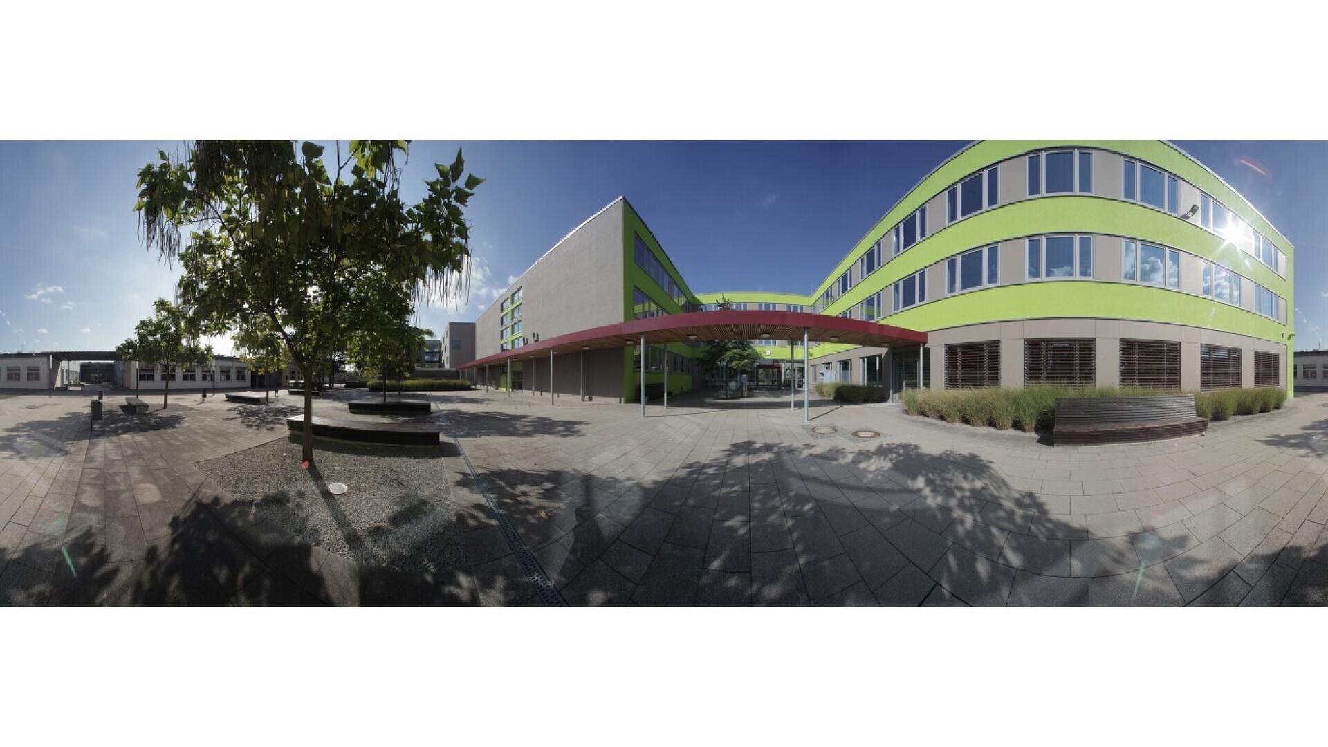 A wide-angle view of a modern school courtyard with benches, trees, and a multi-story building featuring green and yellow horizontal stripes under a clear blue sky.