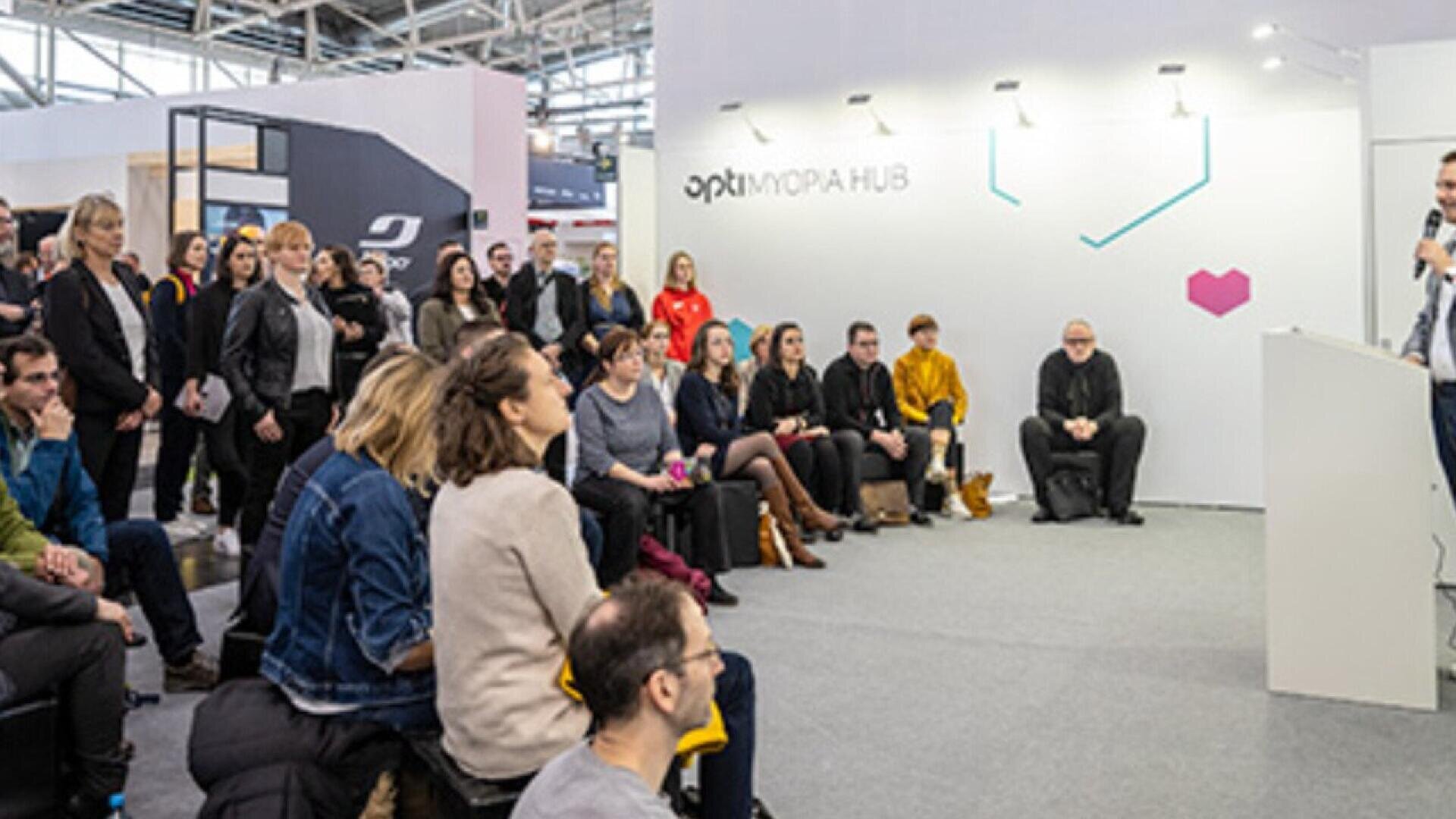 A diverse group of people sits and stands, attentively listening to a speaker at an indoor event or conference in a modern, well-lit space. The wall behind the speaker reads opti MOPA HUB.