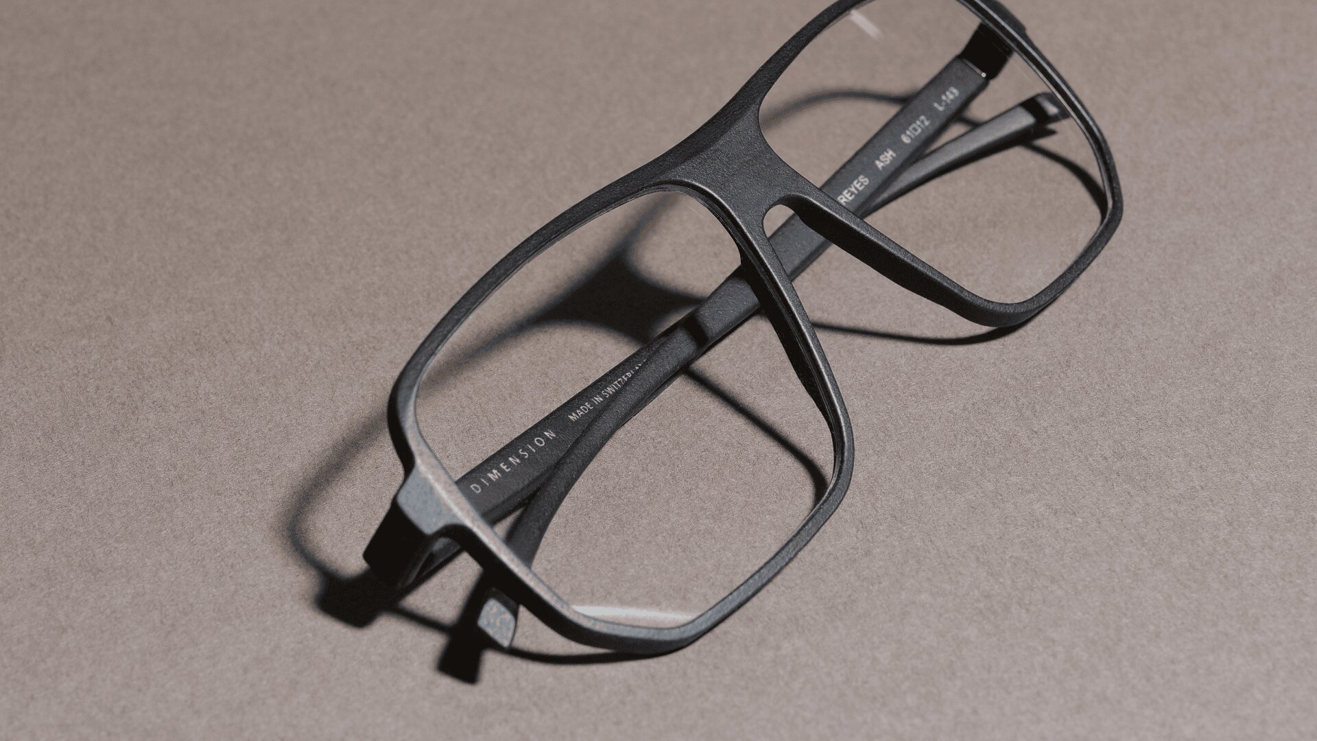 Eine schwarze, rechteckige Brille mit dickem Rahmen ruht auf einer strukturierten grauen Fläche und wirft einen weichen Schatten.