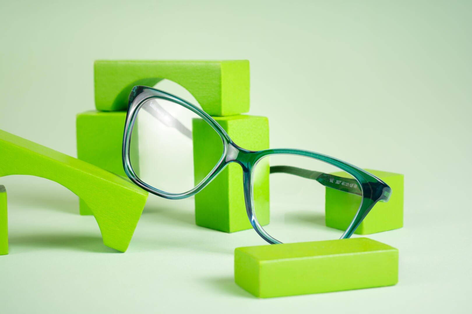 A pair of green-rimmed eyeglasses is displayed among various bright green wooden blocks on a light green background.