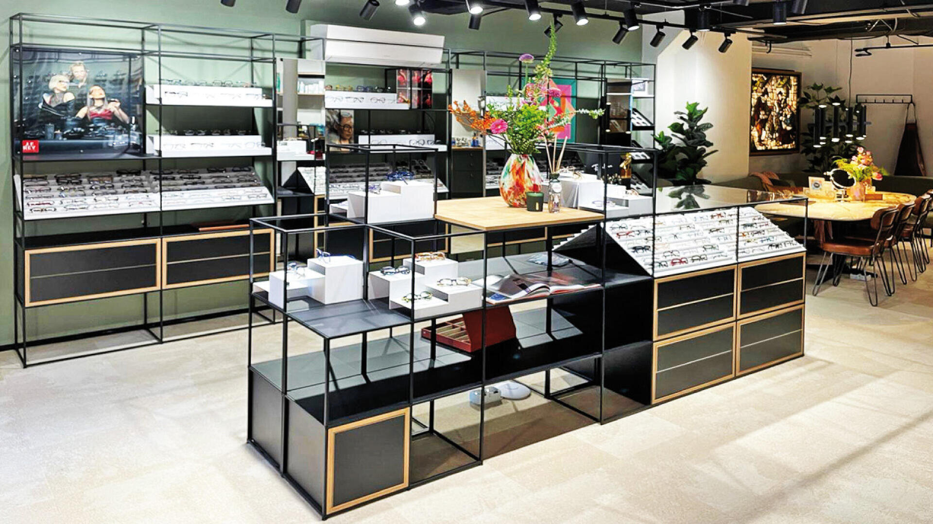 Modernes Interieur eines Brillengeschäfts mit schwarz-goldenen Regalen für verschiedene Brillengestelle, einer zentralen Holztheke mit bunten Blumenarrangements und einem Sitzbereich im Hintergrund. Der Raum ist hell und übersichtlich gestaltet.