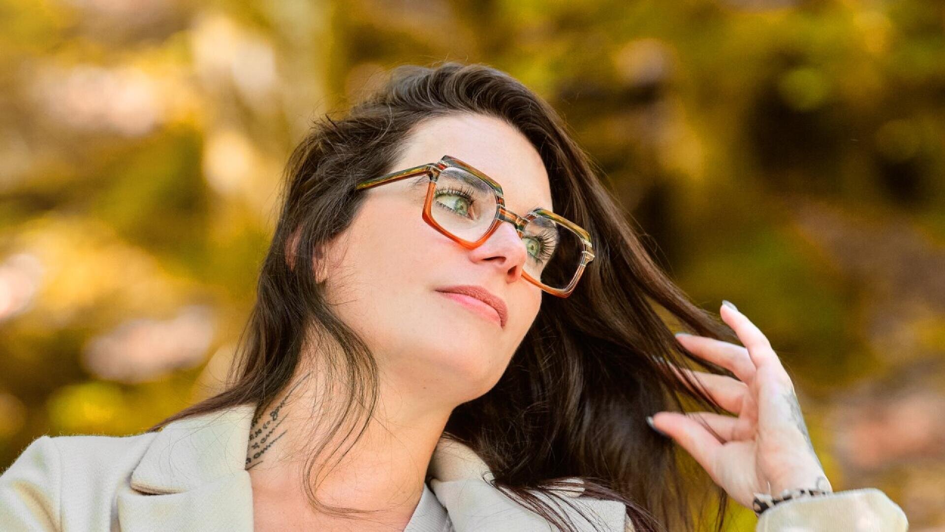 A woman with long dark hair, wearing large square glasses and a light-colored blazer, looks off into the distance while touching her hair. The background is blurred with warm, natural tones.