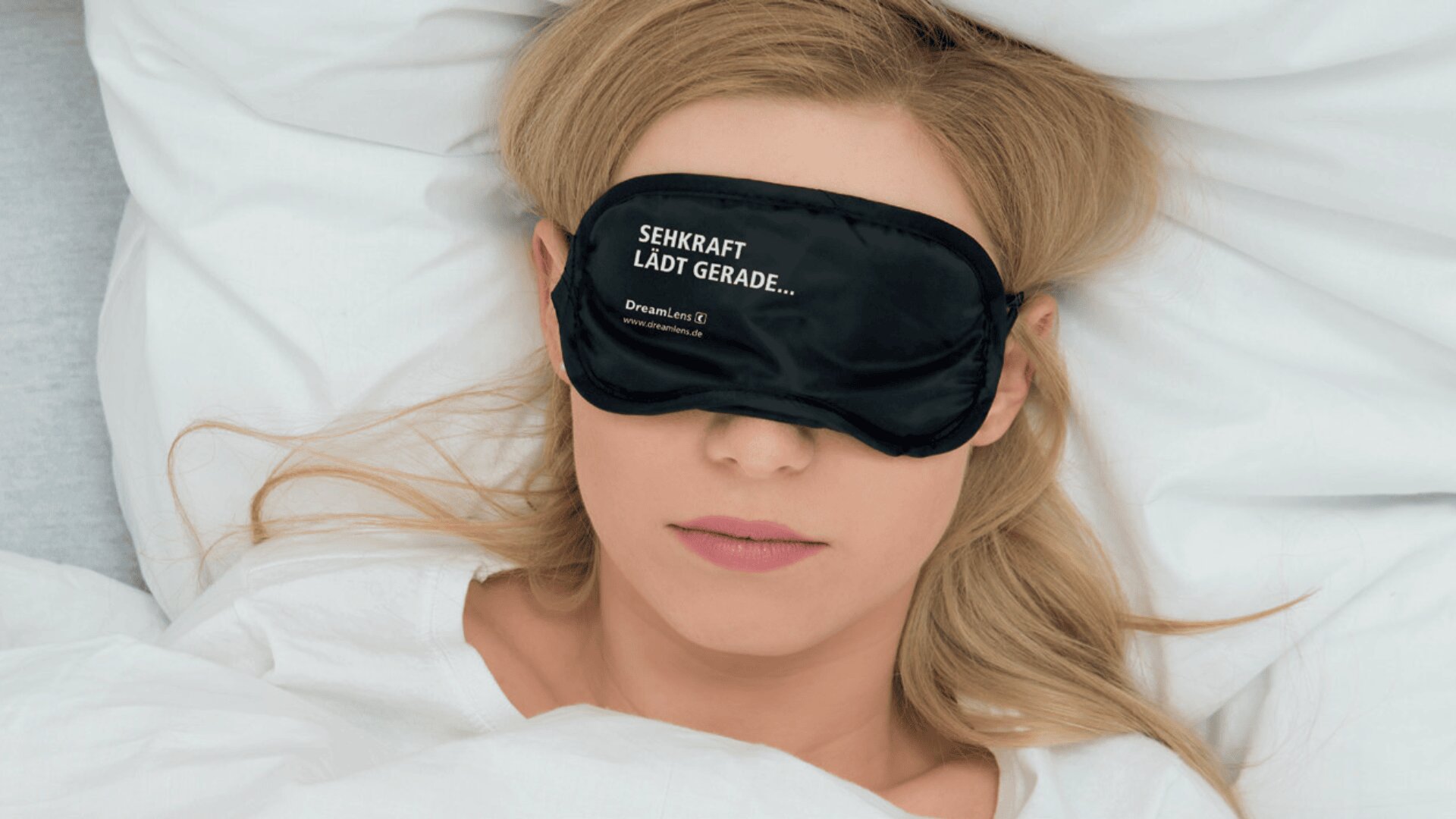 Eine Frau mit blondem Haar liegt im Bett unter einer weißen Decke und trägt eine schwarze Schlafmaske, auf der in weißer Schrift SEHKRAFT LÄDT GERADE... steht.