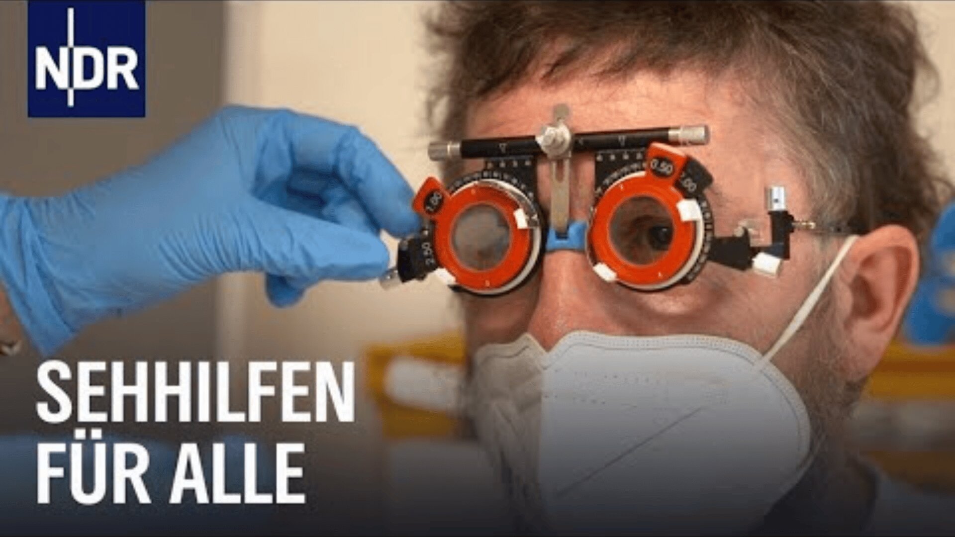 Eine Person, die eine Gesichtsmaske trägt, wird von einer Person mit blauen Handschuhen mit einer Testbrille auf ihr Sehvermögen geprüft. Der Text lautet SEHHILFEN FÜR ALLE und in der linken oberen Ecke befindet sich ein NDR-Logo.