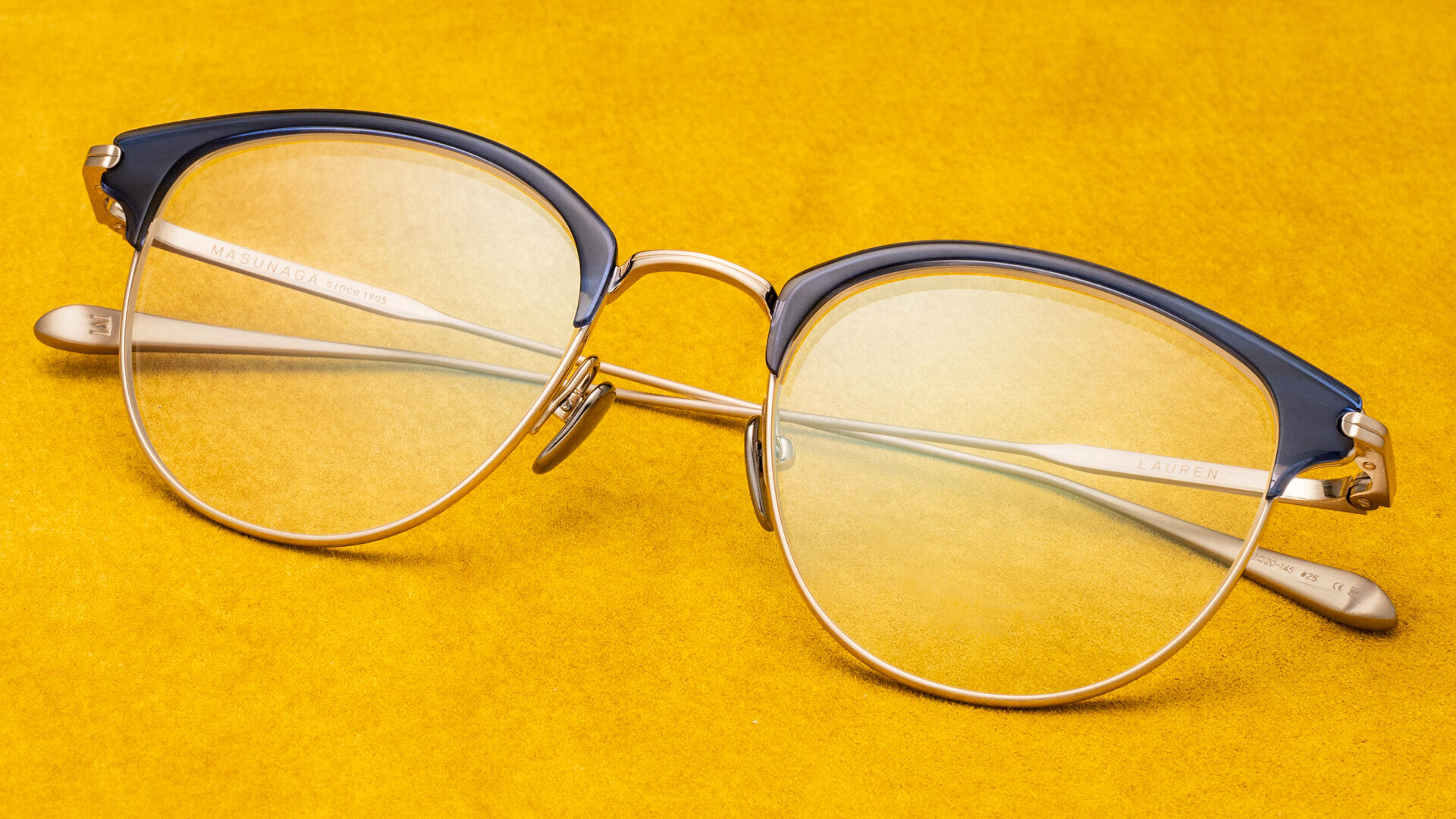 Eine stilvolle Brille mit goldenem Rand und dunkelblauem oberen Rahmen liegt auf einer leuchtend gelben strukturierten Oberfläche.