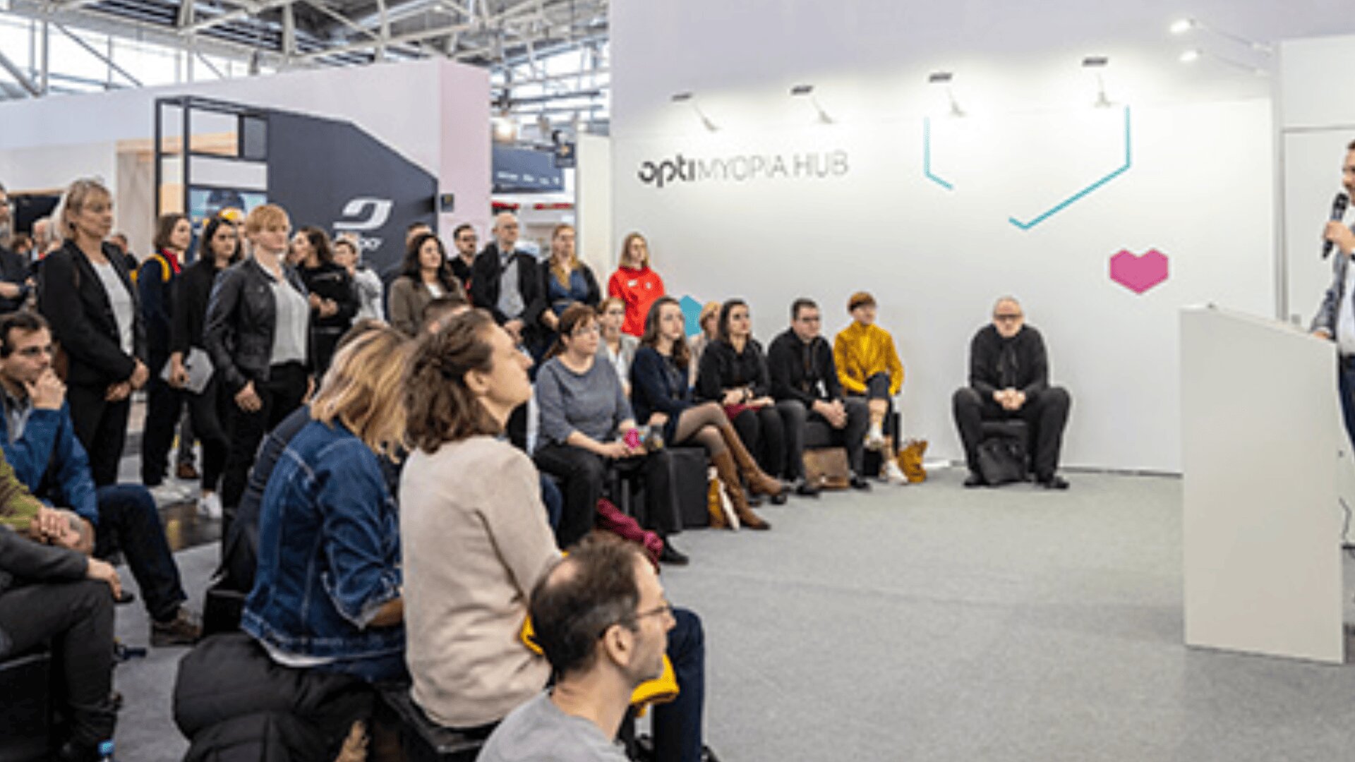 Eine Gruppe von Menschen sitzt und steht aufmerksam und hört einem Redner bei einer Indoor-Veranstaltung zu. Im Hintergrund ist eine weiße Wand mit der Aufschrift OPKMOPAHUB und einer rosa geometrischen Herzform zu sehen.