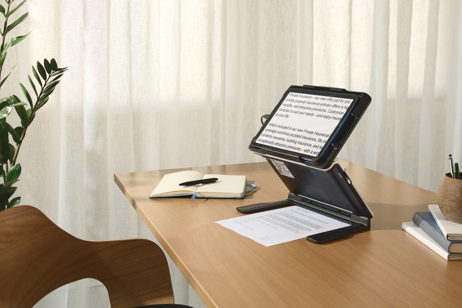 Ein Tablet auf einem Ständer zeigt einen vergrößerten Text auf einem hölzernen Schreibtisch mit Papieren, einem geschlossenen Notizbuch und einem Stift, Büchern und einer Pflanze, neben einem Holzstuhl und vor transparenten Vorhängen.