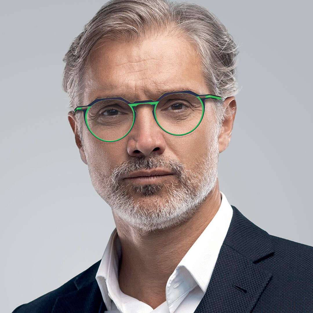 A middle-aged man with gray hair, a short beard, and mustache wears round green glasses and a dark suit, looking seriously at the camera against a plain gray background.