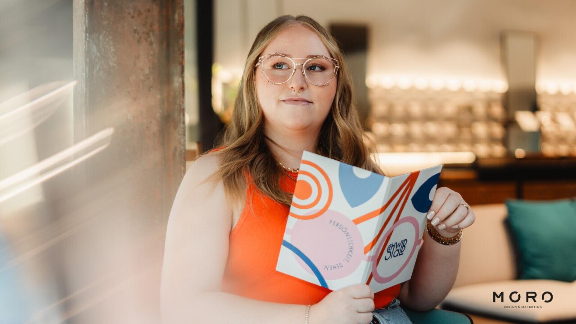 A woman with long, light brown hair and glasses sits indoors, holding a colorful booklet. She wears a sleeveless red top and looks slightly to the side. The background is softly blurred. The logo MGRO is visible in the corner.