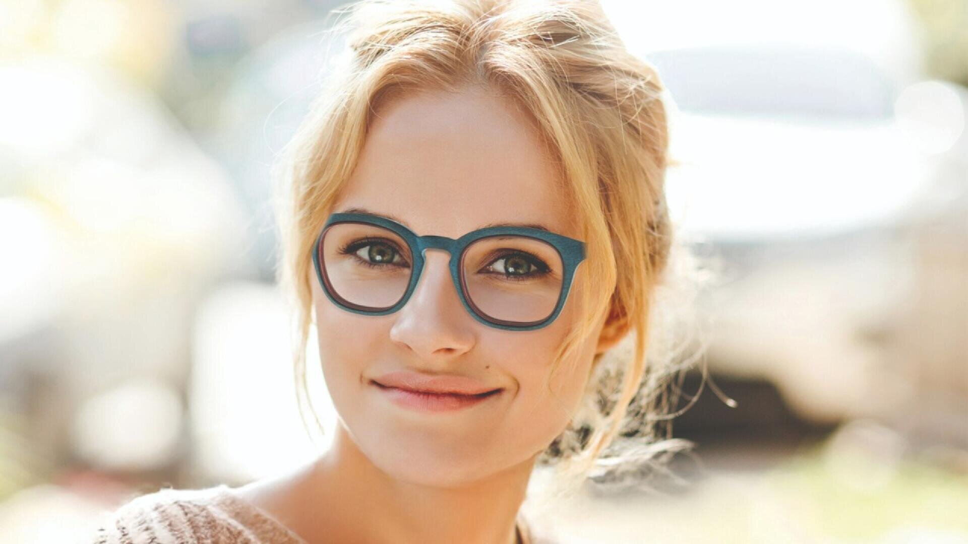 A young woman with blonde hair and blue glasses smiles gently while looking at the camera. The background is blurred and bright, suggesting an outdoor setting.