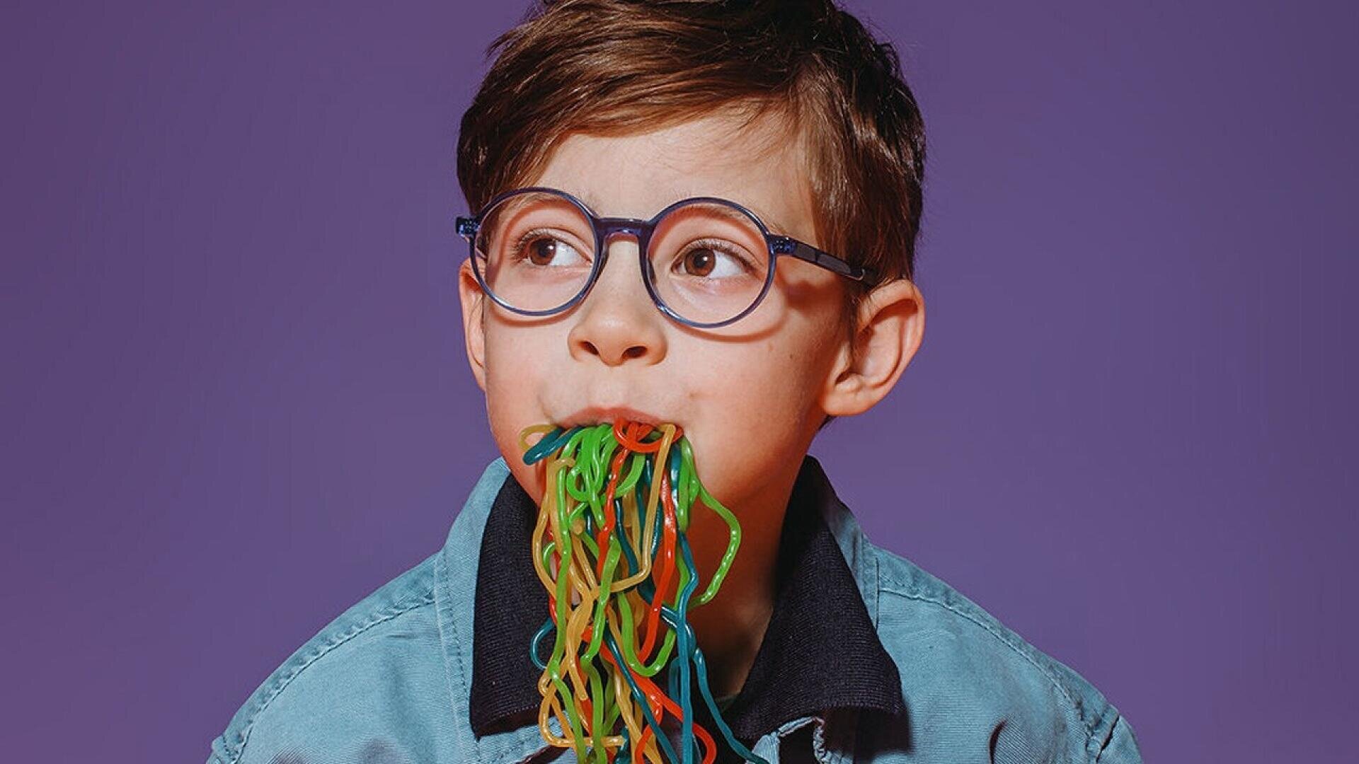 A young boy with glasses and a denim jacket has colorful candy strings hanging from his mouth against a purple background.