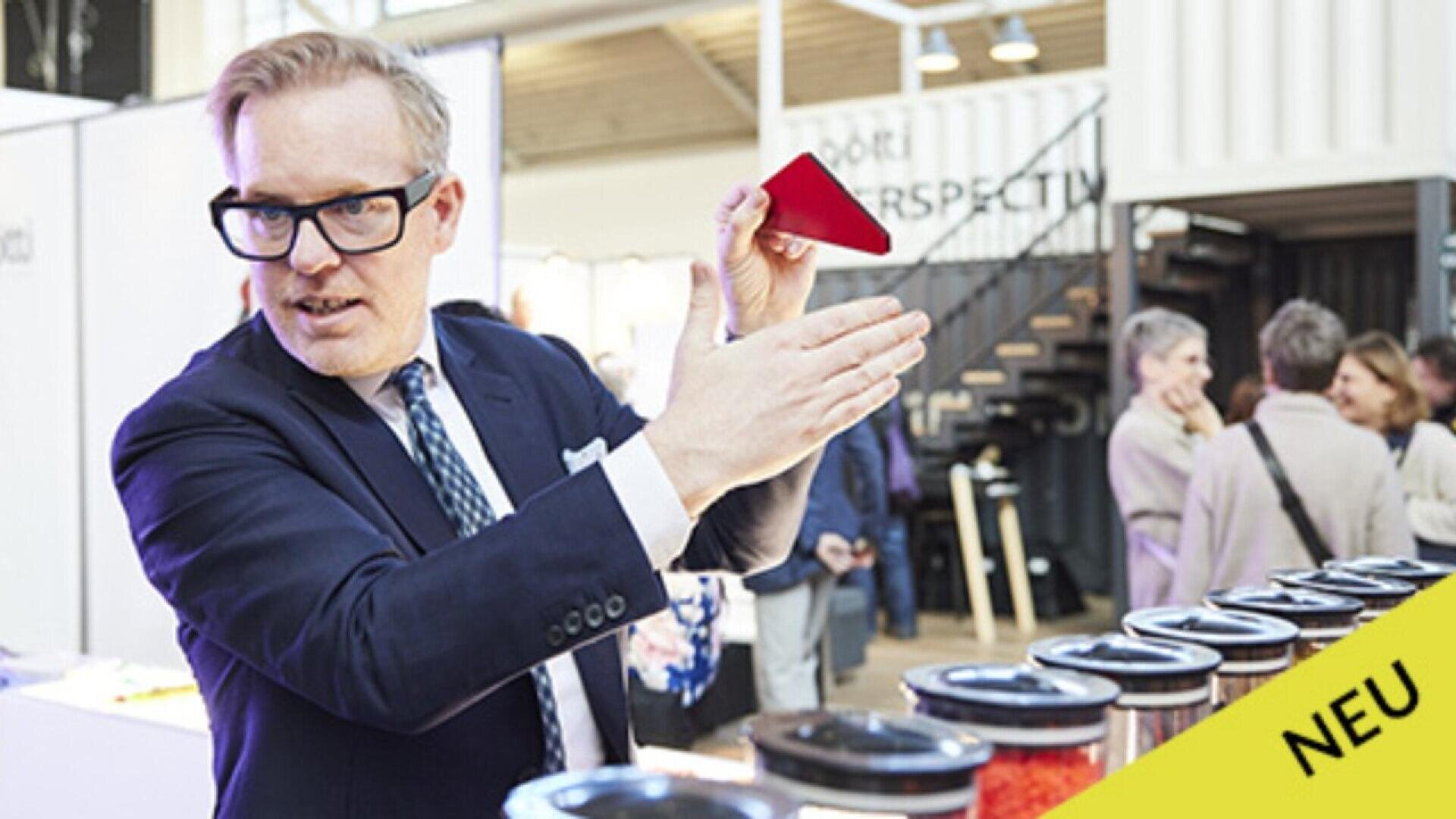 A man in a suit and glasses gestures with a red object in his hand at an indoor event with people in the background. Several containers are lined up on a table in front of him. A yellow label reads NEU.