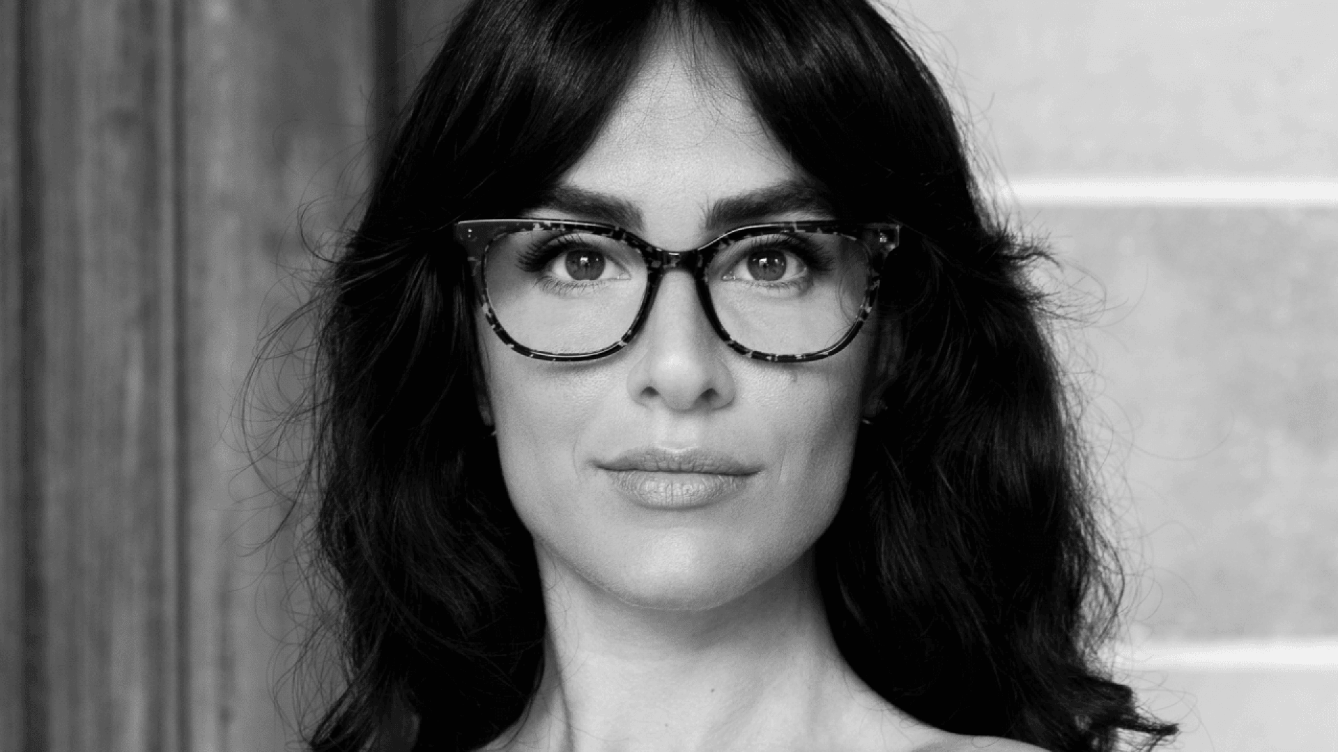 A woman with dark wavy hair and glasses looks directly at the camera with a neutral expression. The image is in black and white, and the background is softly blurred.