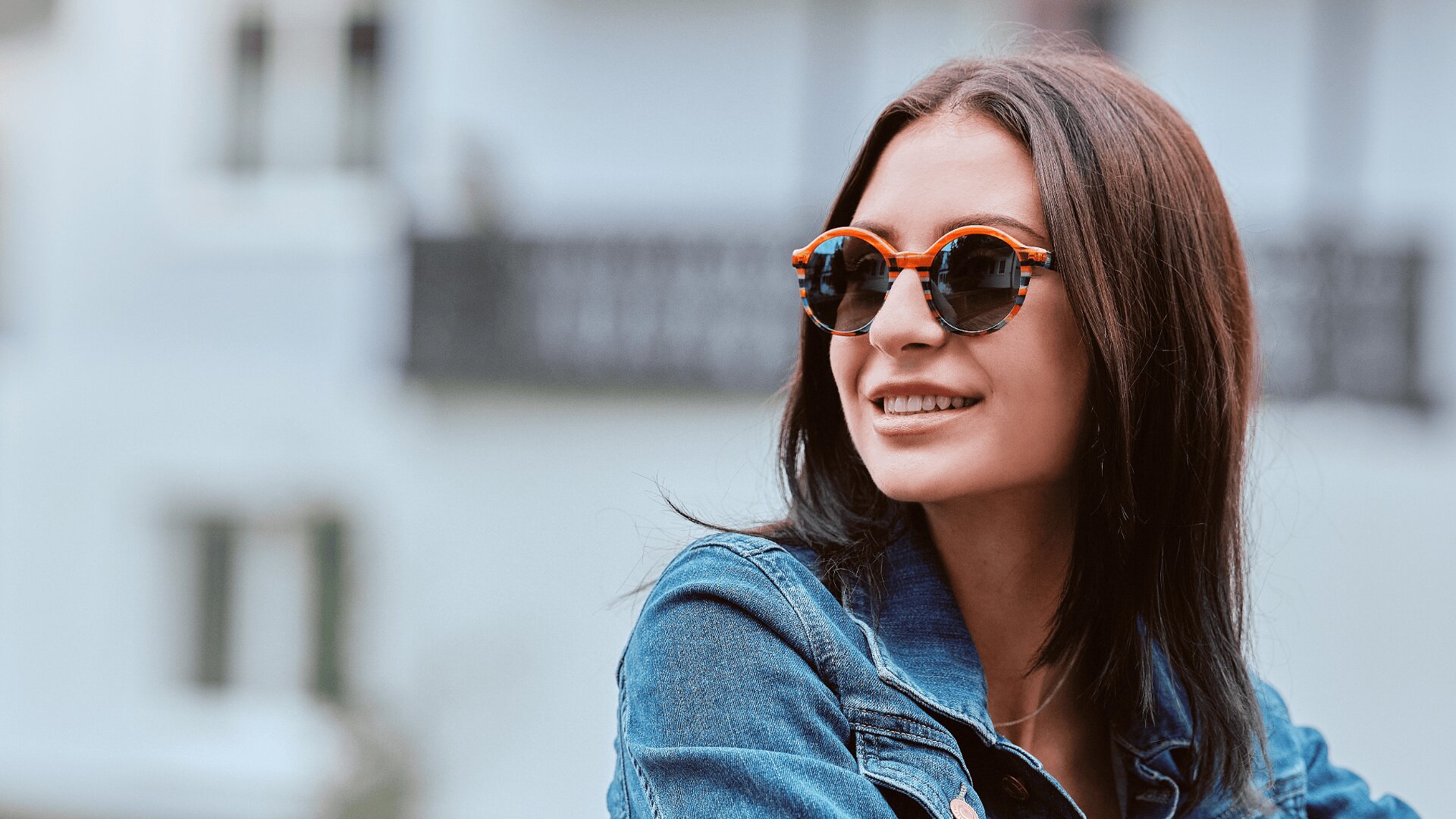 Eine junge Frau mit glattem braunem Haar lächelt, während sie eine runde Sonnenbrille und eine Jeansjacke trägt. Sie steht im Freien mit unscharfen Gebäuden im Hintergrund.
