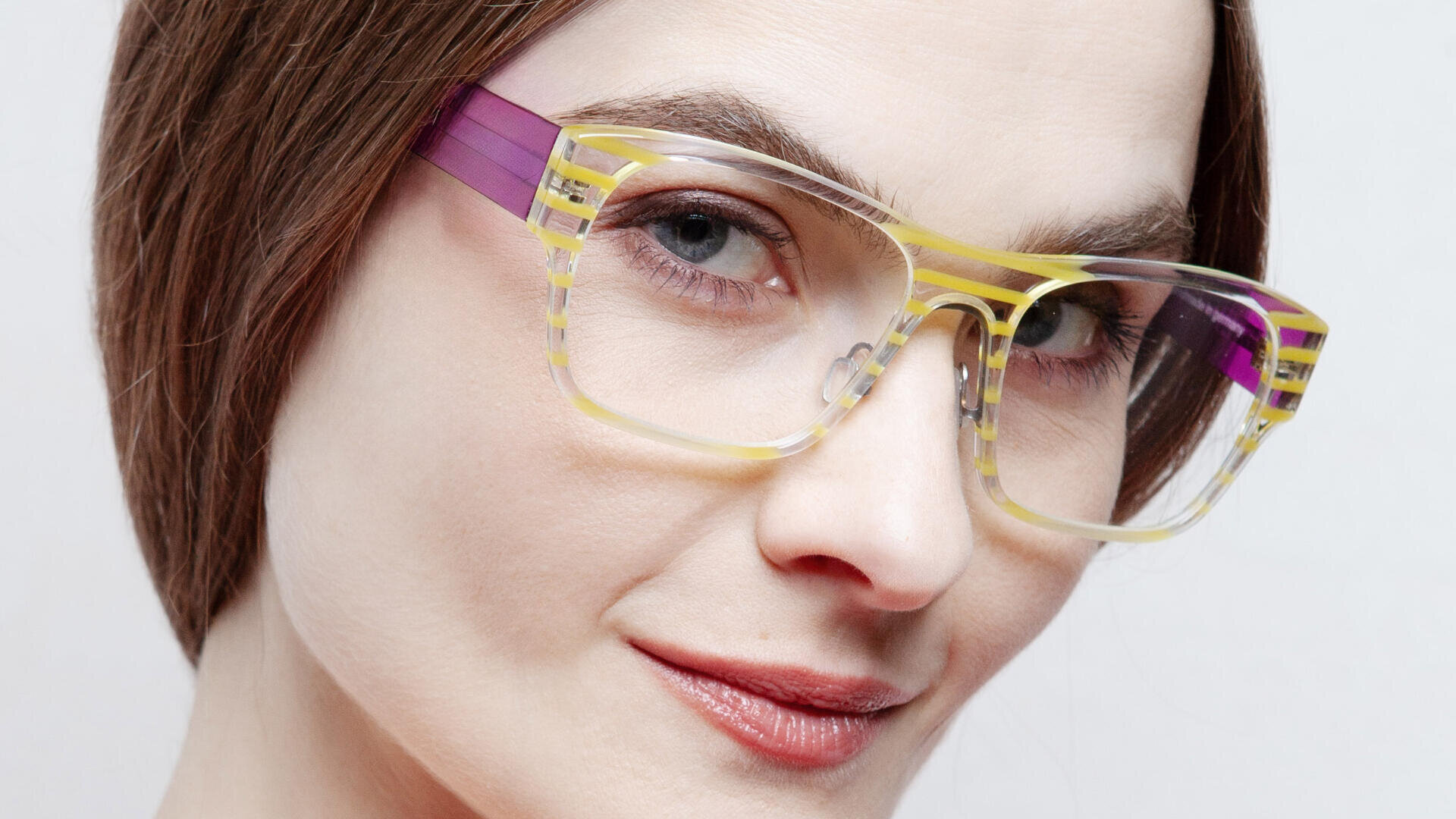 Eine Frau mit heller Haut und braunem Haar blickt in die Kamera. Sie trägt eine auffällige Brille mit transparentem Rahmen in Gelb und Lila. Sie lächelt leicht und hinter ihr befindet sich ein neutraler Hintergrund.