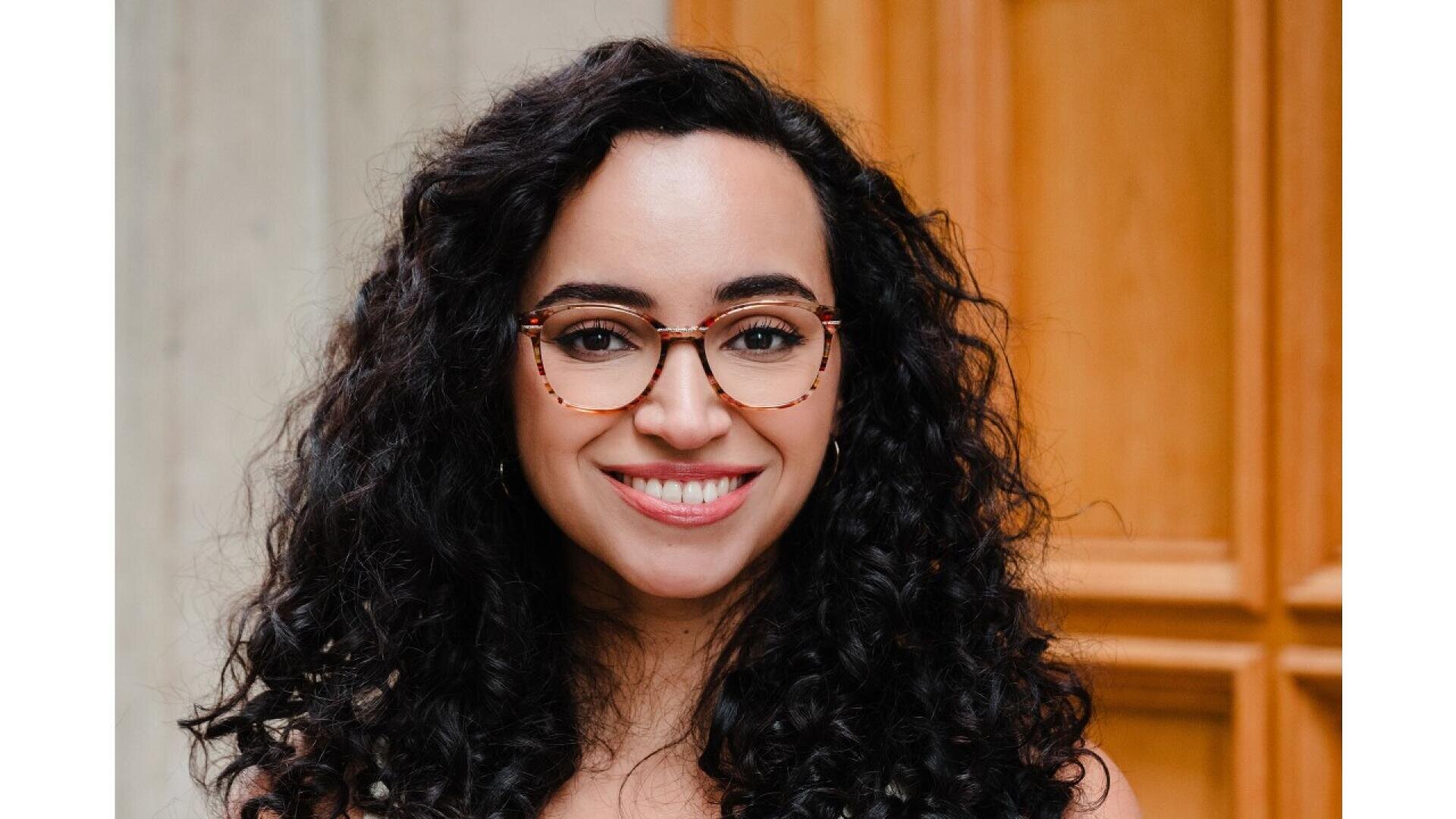 A woman with long, curly dark hair and glasses is smiling. She is standing in front of a blurred wooden and light-colored wall background.