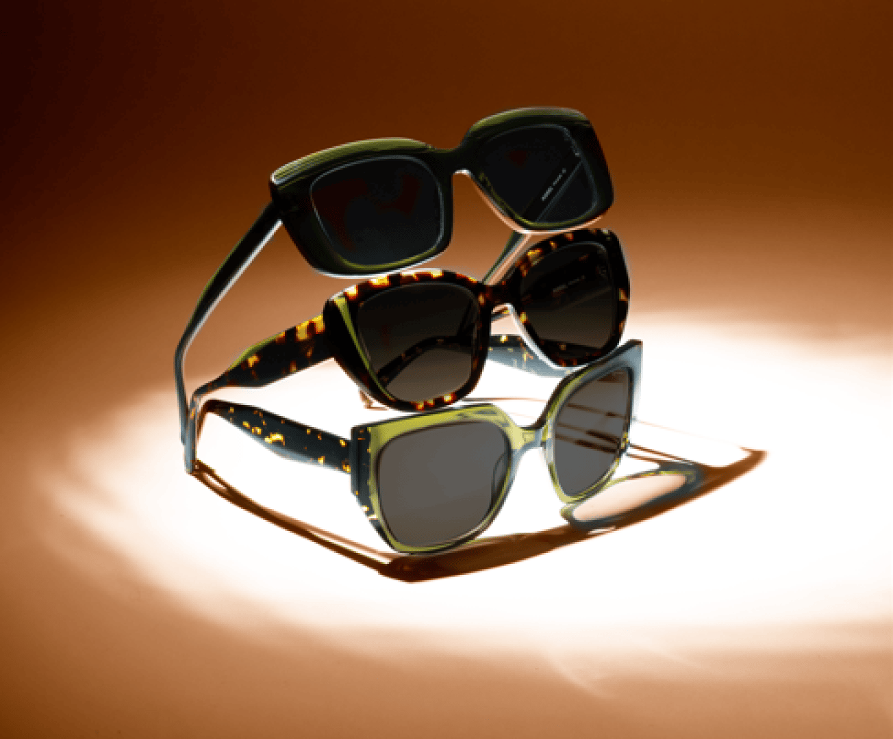 Three pairs of stylish sunglasses are stacked vertically on a brown background, each with thick frames in different colors and patterns, including solid green, tortoiseshell and speckled green.