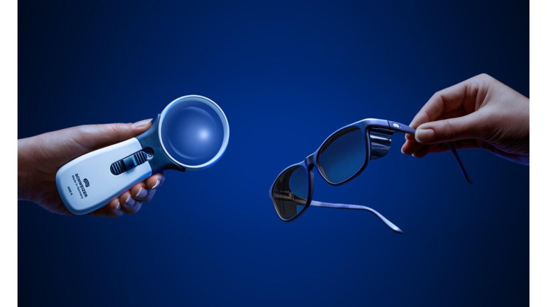 A hand holds a magnifying glass with a built-in light, facing another hand holding a pair of dark sunglasses. The objects are set against a deep blue background.