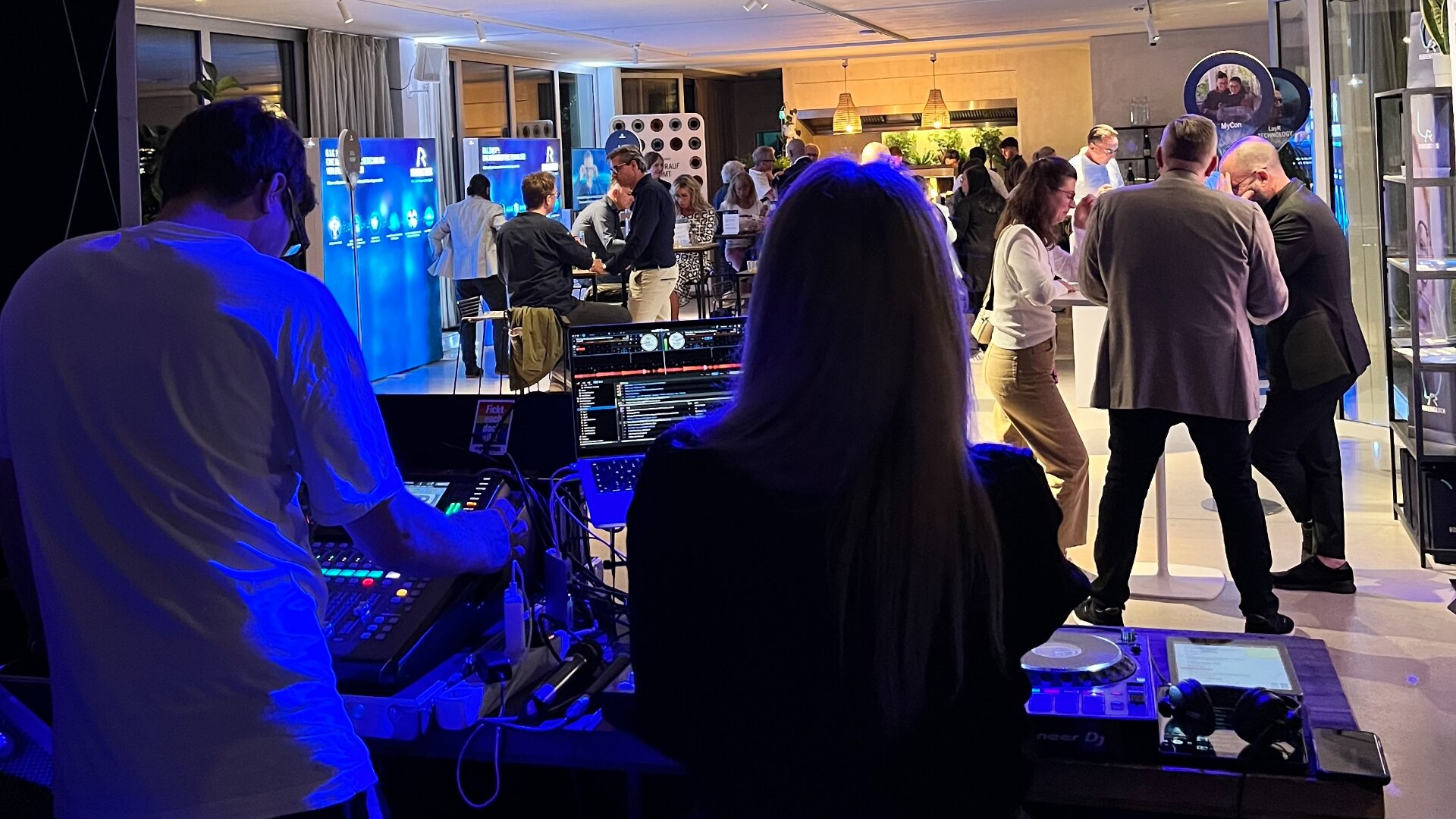 Ein DJ und ein Tontechniker bedienen die Geräte bei einer Indoor-Veranstaltung, bei der sich die Leute in der Nähe der Stände bei warmer und blauer Beleuchtung austauschen.