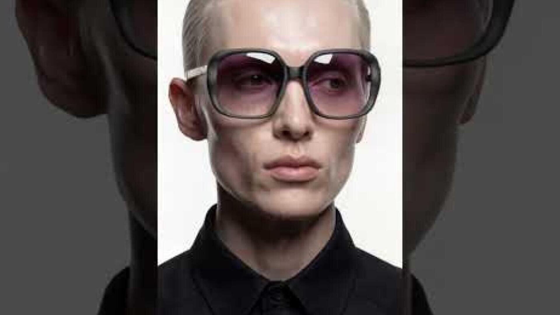 A person with short blond hair wears large, dark sunglasses and a black collared shirt and looks slightly to the side against a plain, light-colored background.