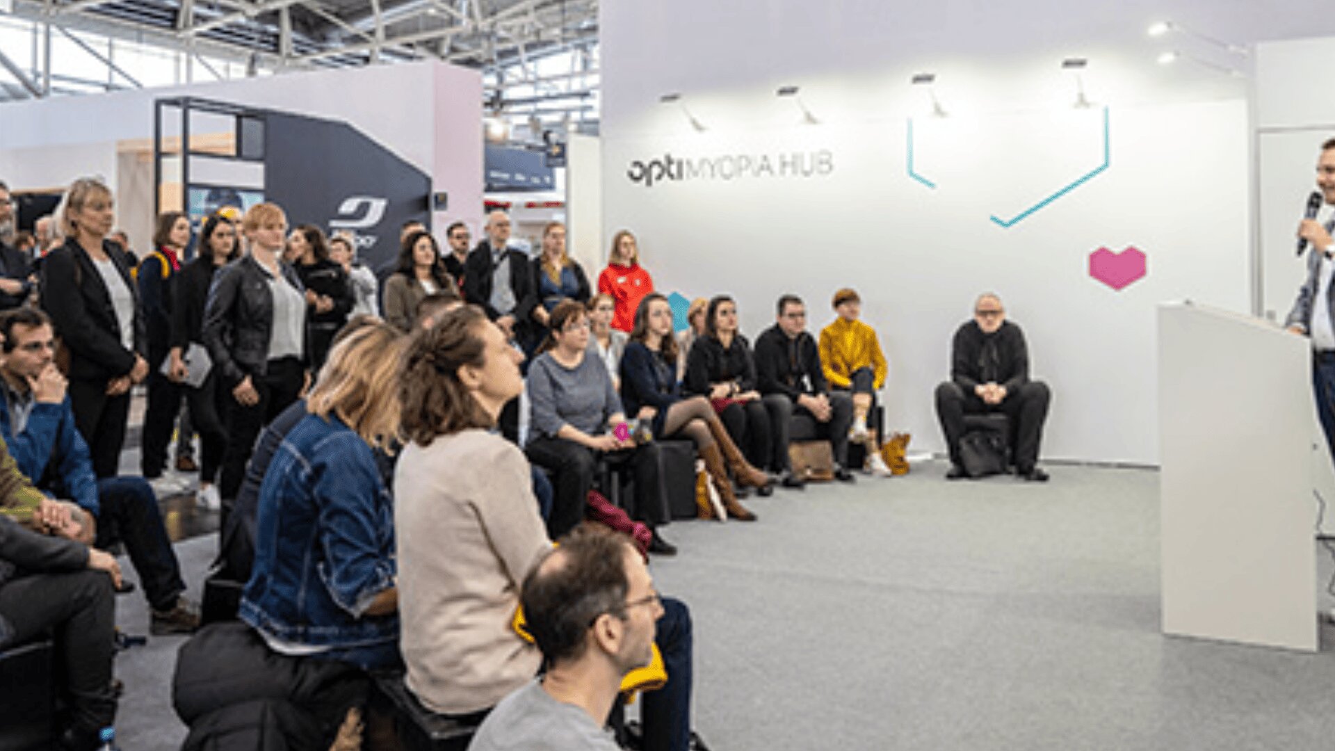 Eine Gruppe von Menschen sitzt und steht aufmerksam da und hört einem Redner bei einer Veranstaltung oder Konferenz in einer Halle zu, in der Nähe einer weißen Wand mit OPTIMORAPHUB und einem kleinen rosa Herz-Logo.